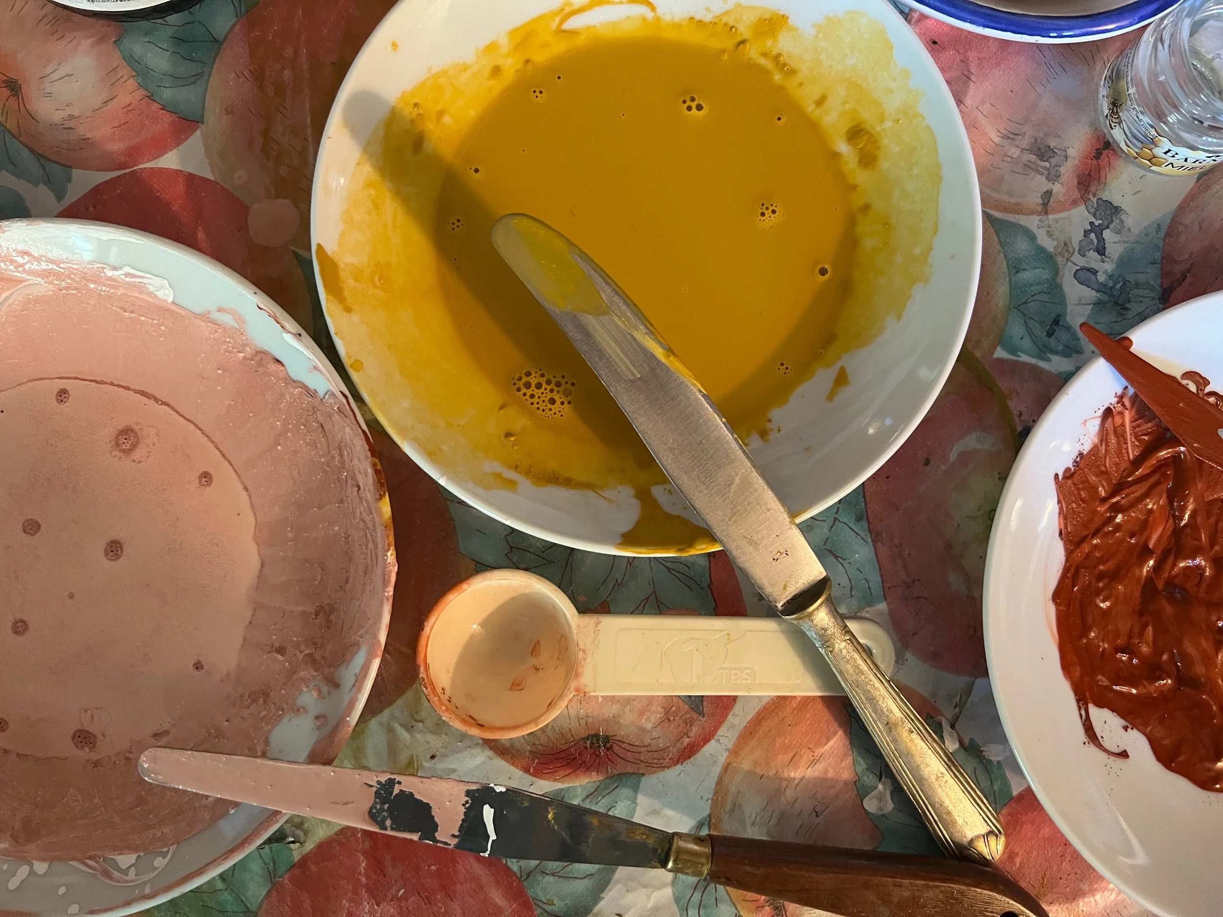 Bowl of yellow batter, bowl of pink batter, bowl of chocolate batter, plastic spatula, knife, small empty bowl, on a colorful apple-patterned tablecloth.