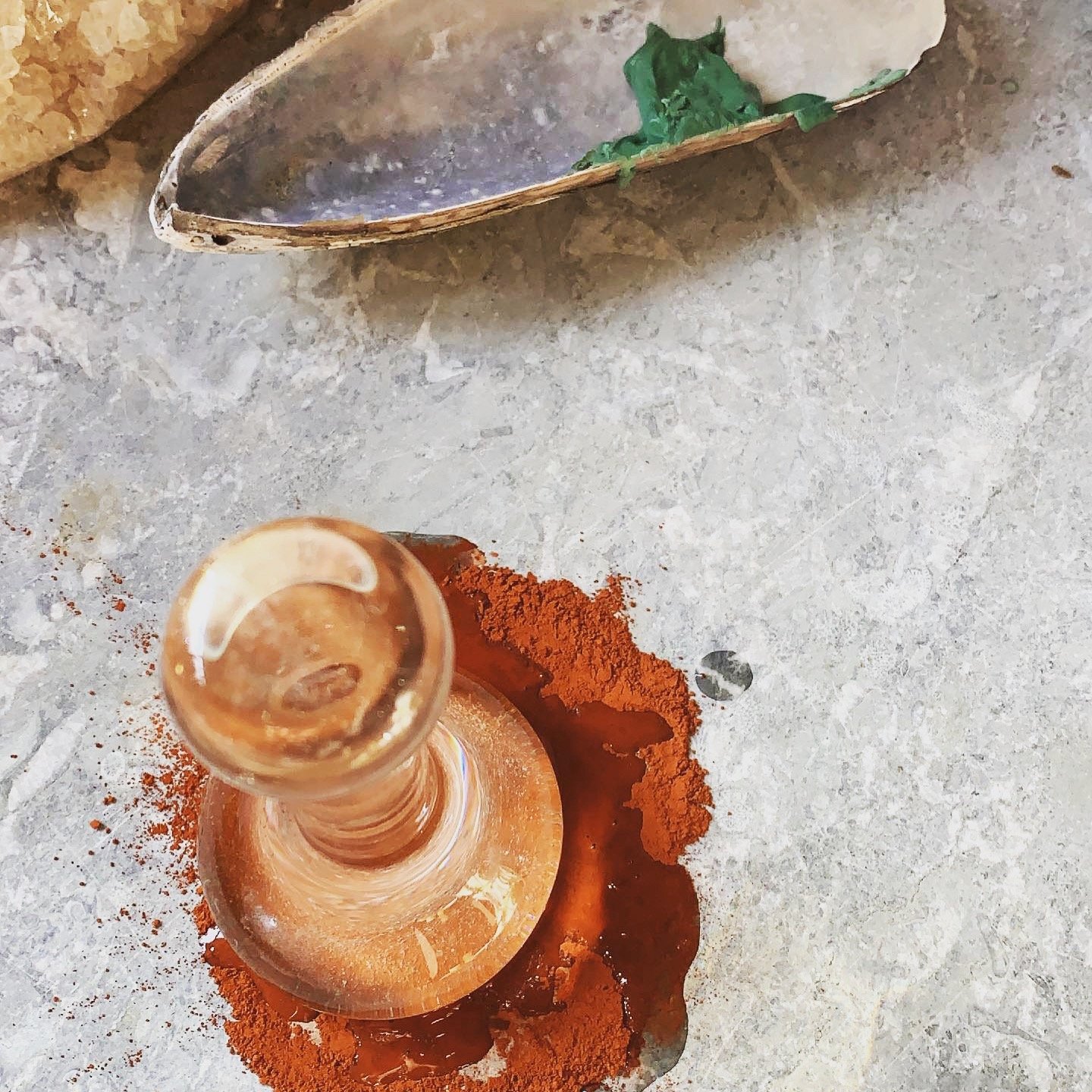 A top-down view of a glass candle holder with a lit candle inside, surrounded by red chili powder on a gray concrete surface. In the background, there is a spoon with some green wax and a piece of wax near the top of the image.
