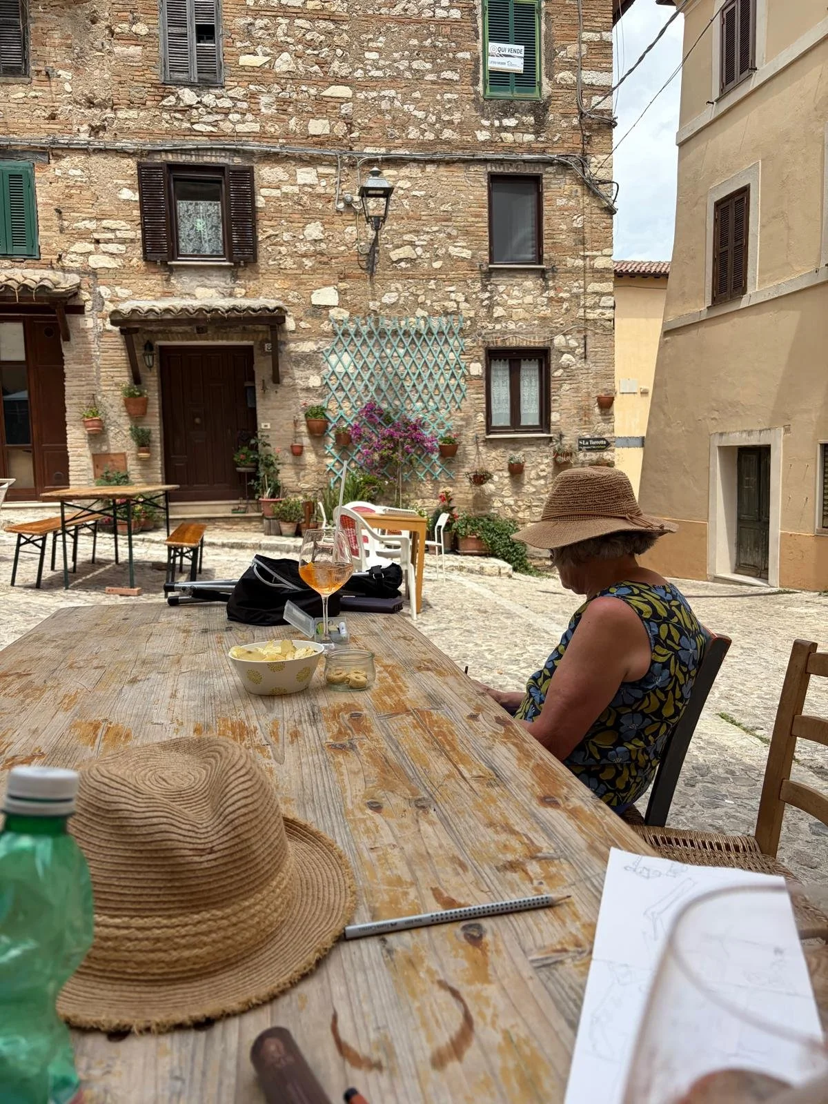 Woman sitting at an outdoor table on a cobblestone street, wearing a wide-brimmed hat and a floral sleeveless dress, with items like a hat, water bottle, and papers on the table, in a rustic European village setting with old stone buildings and plant