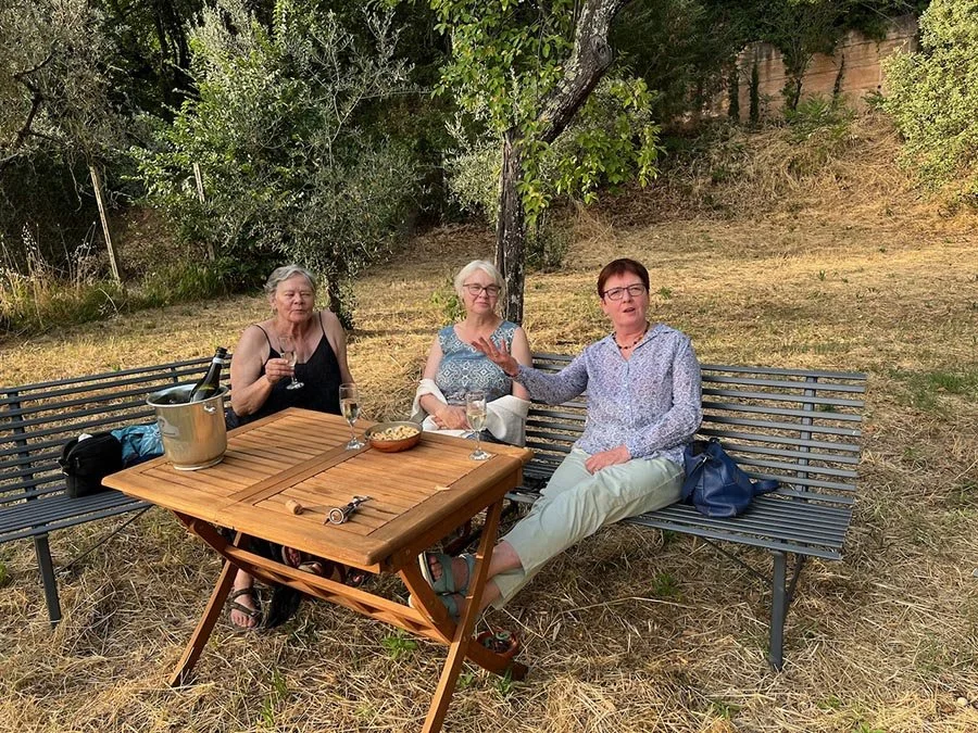 Three women sitting on a metal bench outdoors under a tree, enjoying drinks and snacks at a wooden table.