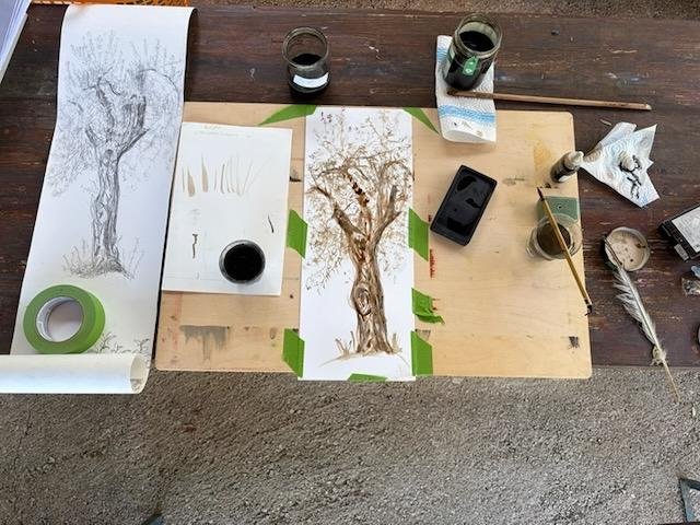 Art supplies and sketches of a tree on a workspace, including paint jars, brushes, tape, and a smartphone.