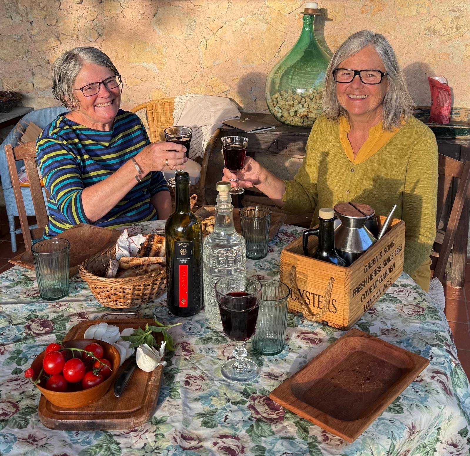 Two older women with gray hair and glasses smiling and raising glasses of red wine at a dining table outdoors. The table has wine bottles, glasses, a basket of bread, and a wooden block with condiments. A large decorative glass bottle and stone wall 