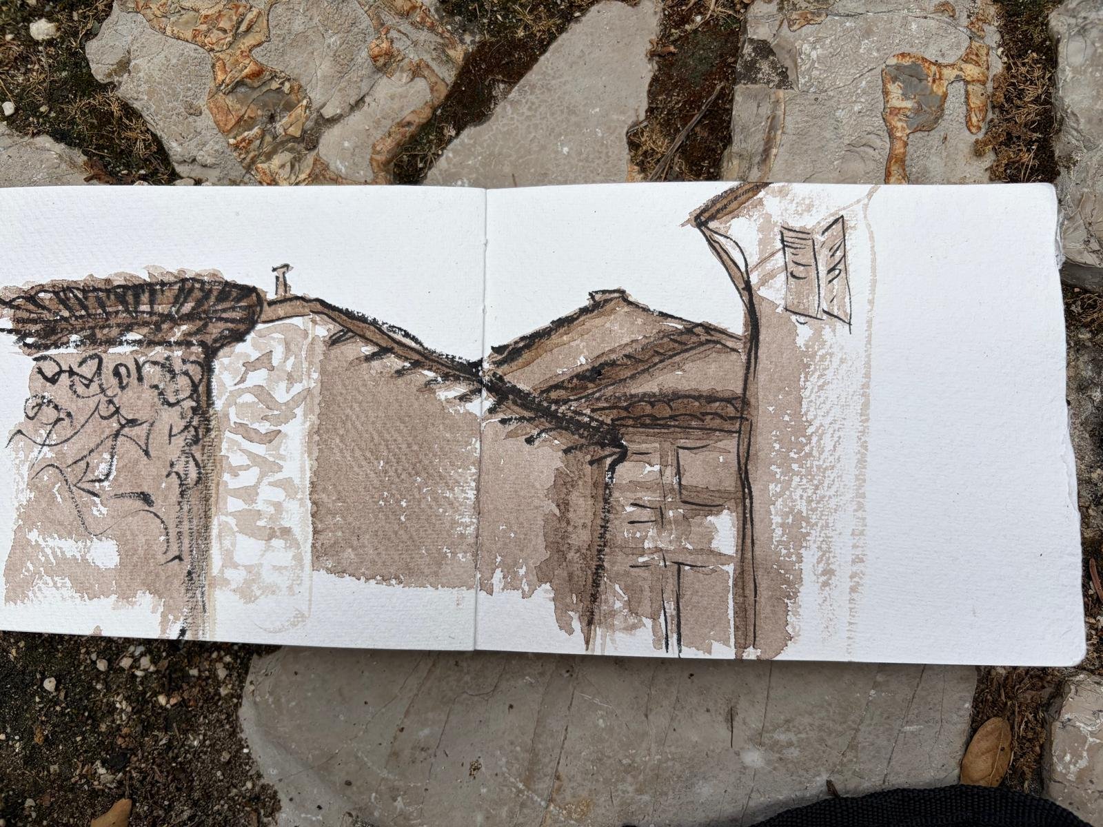 A sketch of a house with a sloped roof, a chimney, and a window, drawn in brown ink on a white sketchpad resting on a rocky outdoor surface from Charlotte Andrew Art Retreat in the Sabine Hills, Italy.