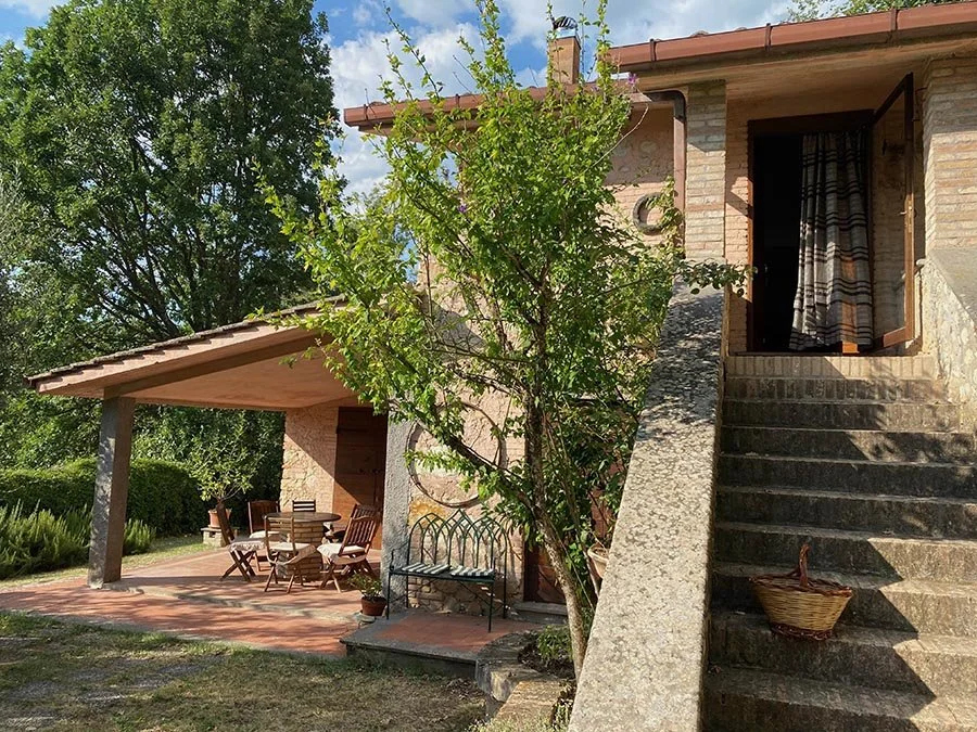 A stunning brick house with outdoor stairs leading to an open door and small patio with a dining table and chairs is under a roof extension, surrounded by greenery and trees for Art at Sabine Hills retreat