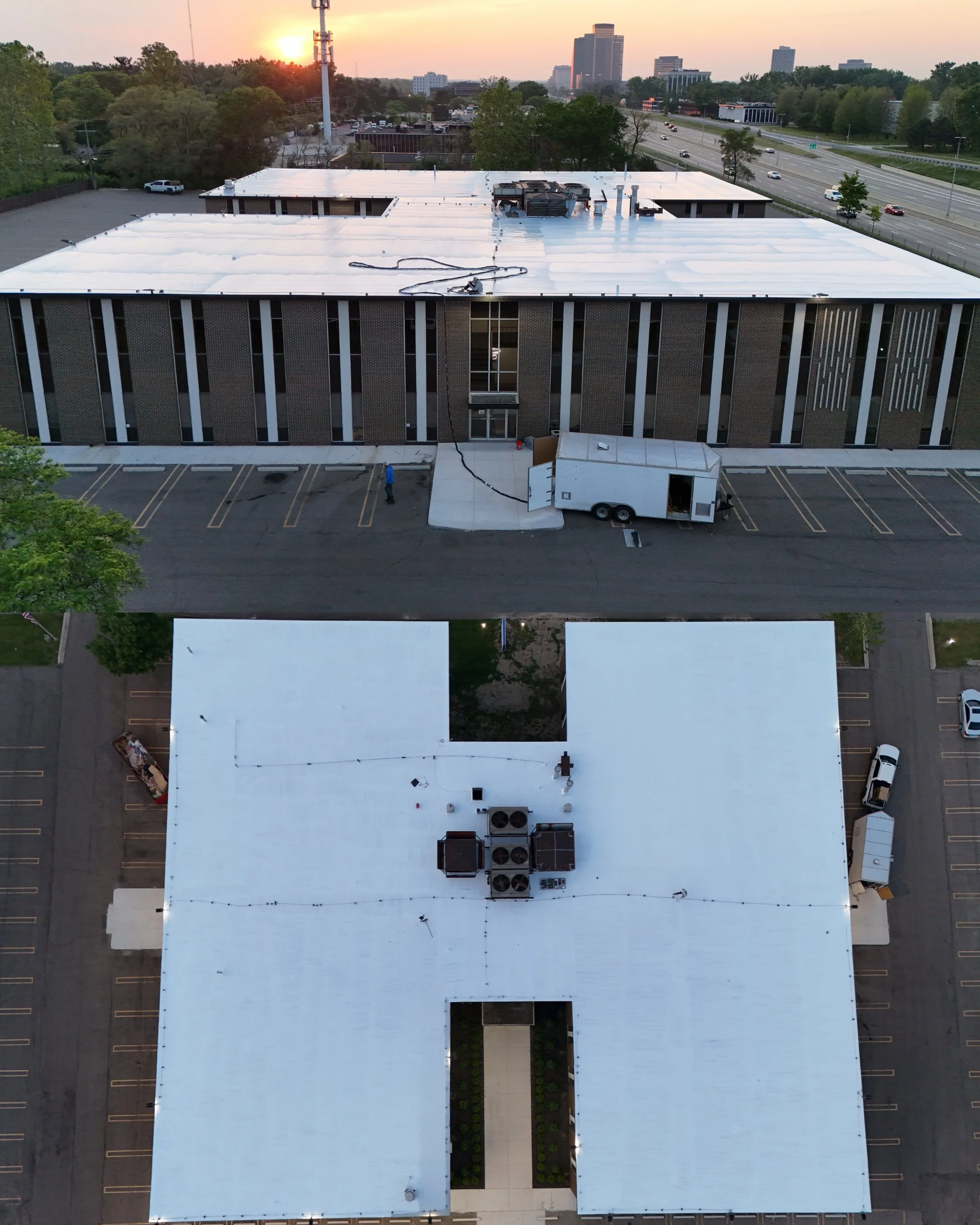 An aerial view of a large building with a white roof, a parking lot, and surrounding trees at sunset. A maintenance trailer is parked in the lot.
