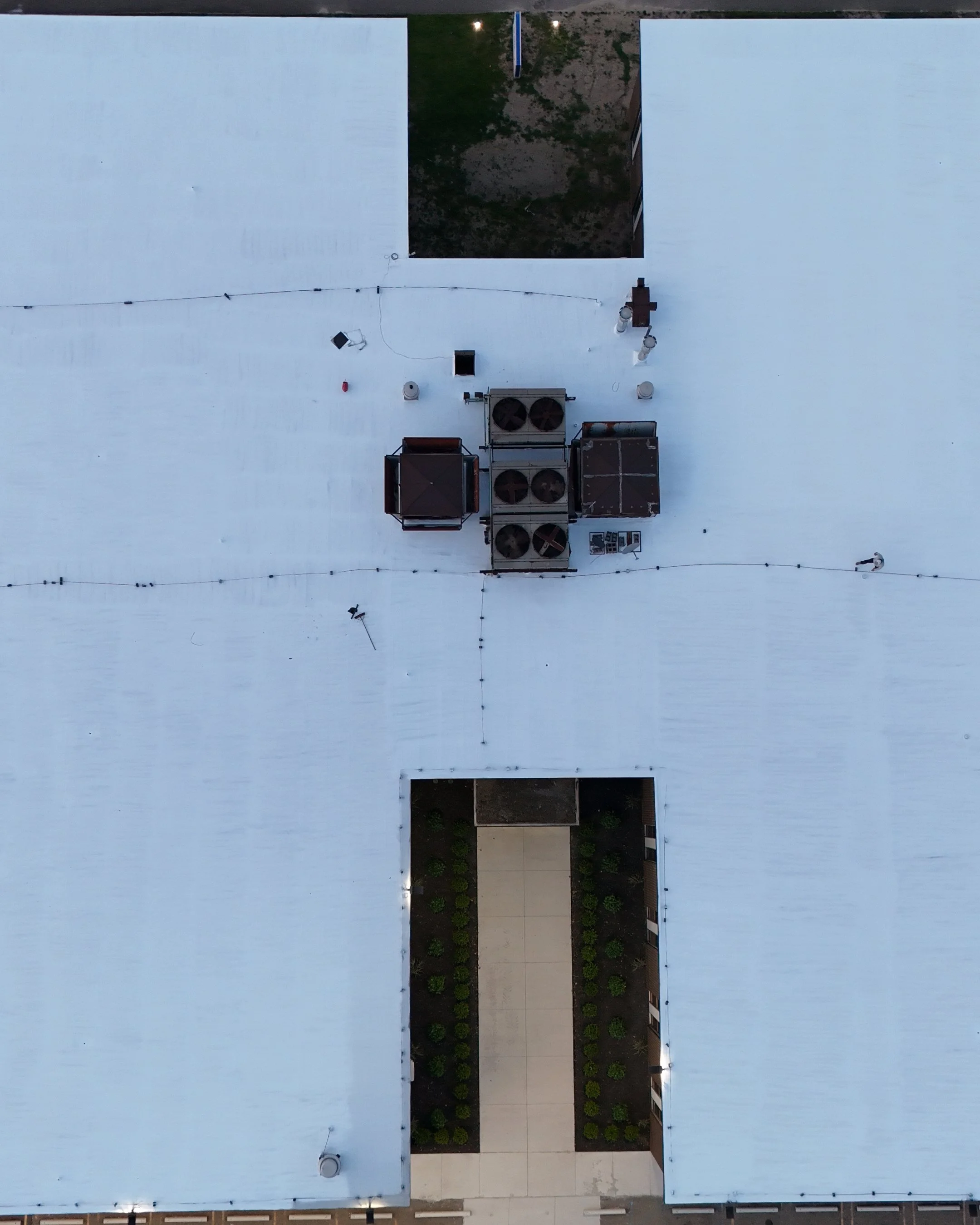 Aerial view of a building rooftop with HVAC units, vents, and string lights, with a pathway and small garden area visible at the bottom.