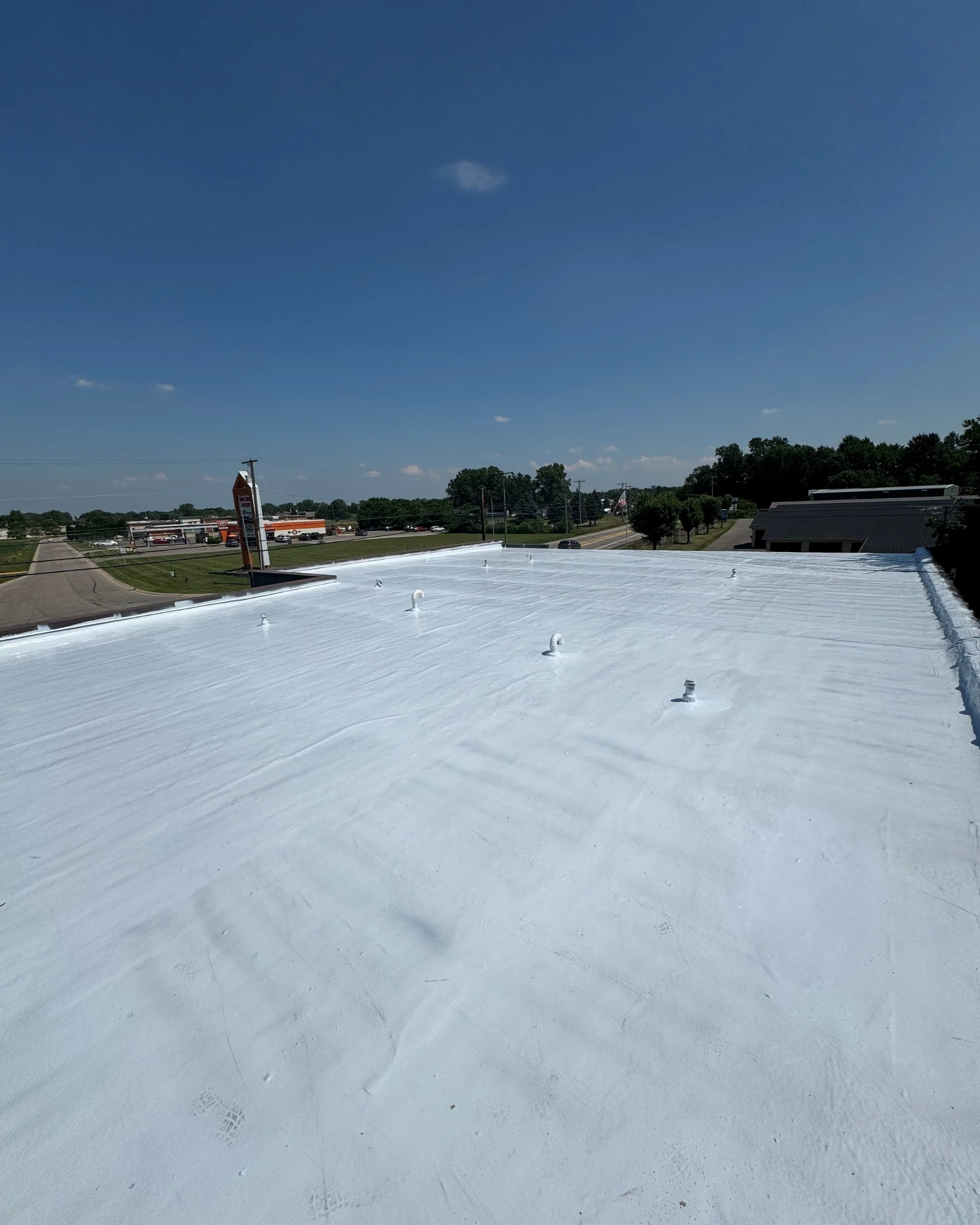 White commercial building roof with several vent pipes and a bright blue sky with some clouds in the background.