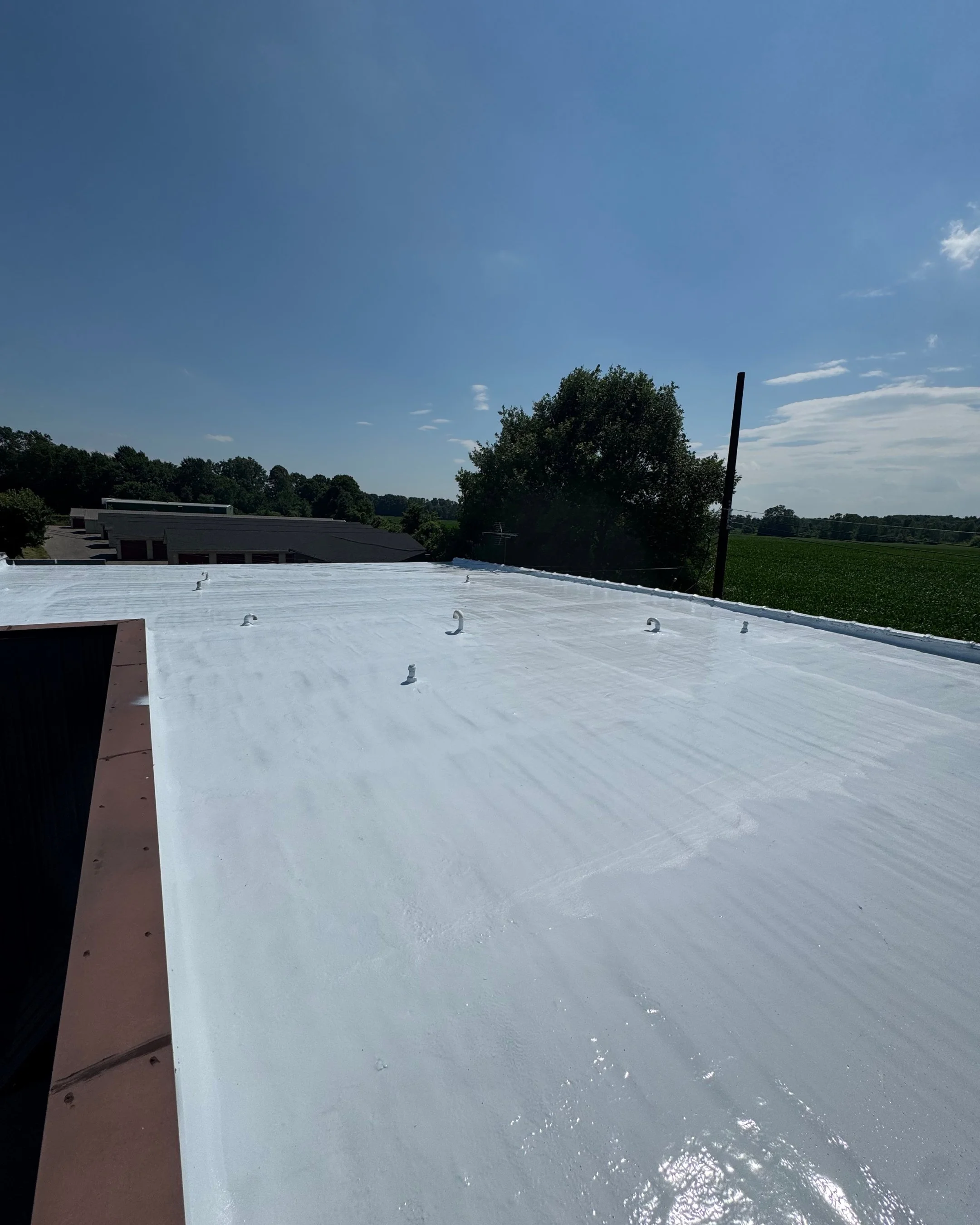 White commercial building roof with raised roof hooks, under a bright blue sky, trees and open fields in the background.