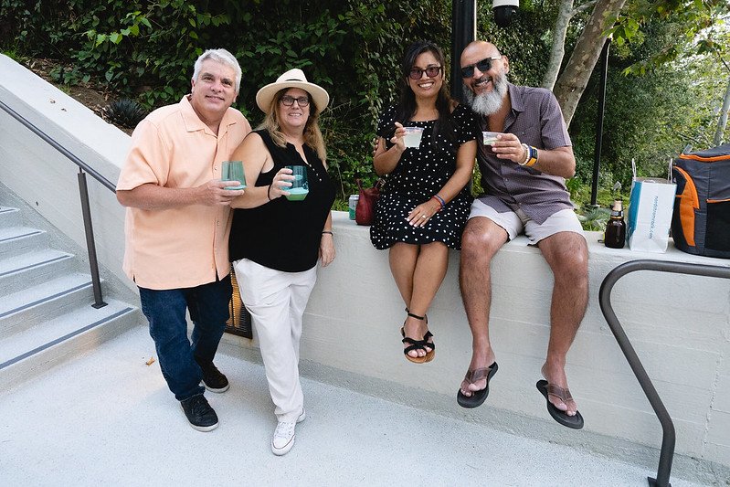 Four friends enjoying drinks outdoors, with two standing and two sitting on a low wall, surrounded by greenery.