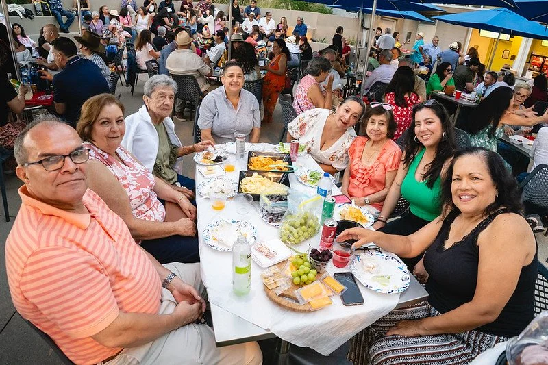 A group of nine people sitting around a table outdoors, enjoying food and drinks at a social gathering, with other groups dining in the background.