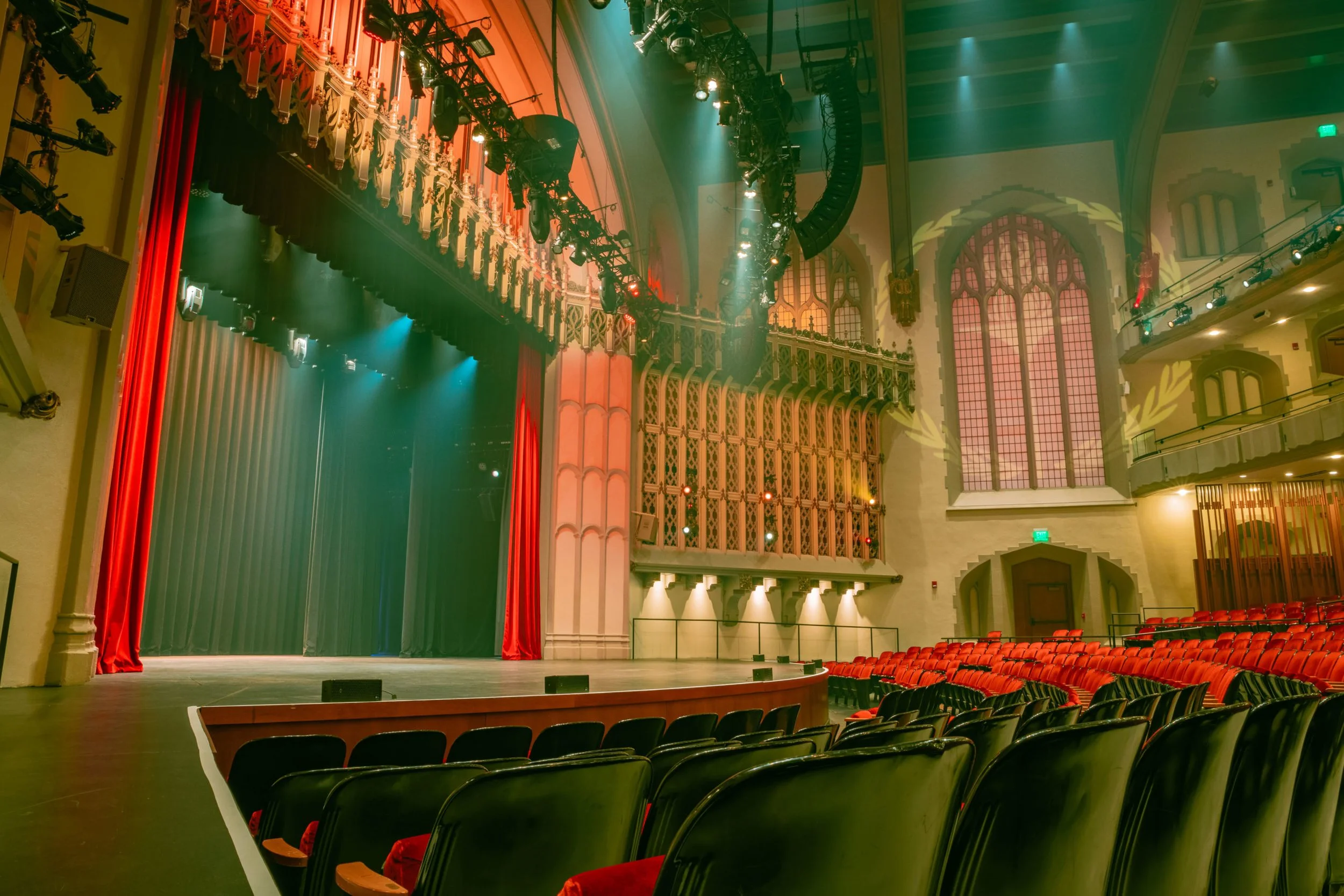 Empty theater stage with closed green curtain, red velvet side curtains, and an ornate stage front with sconces. Audience seating with red and black chairs in a large, well-lit auditorium featuring stained glass windows.