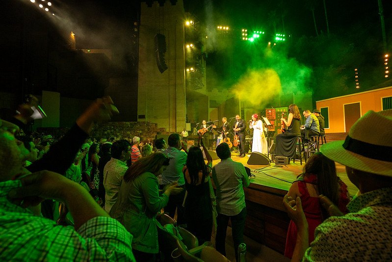 Live music performance on stage with multiple musicians playing instruments and singing, audience watching and enjoying the concert at an indoor venue with colorful lights and stage fog.