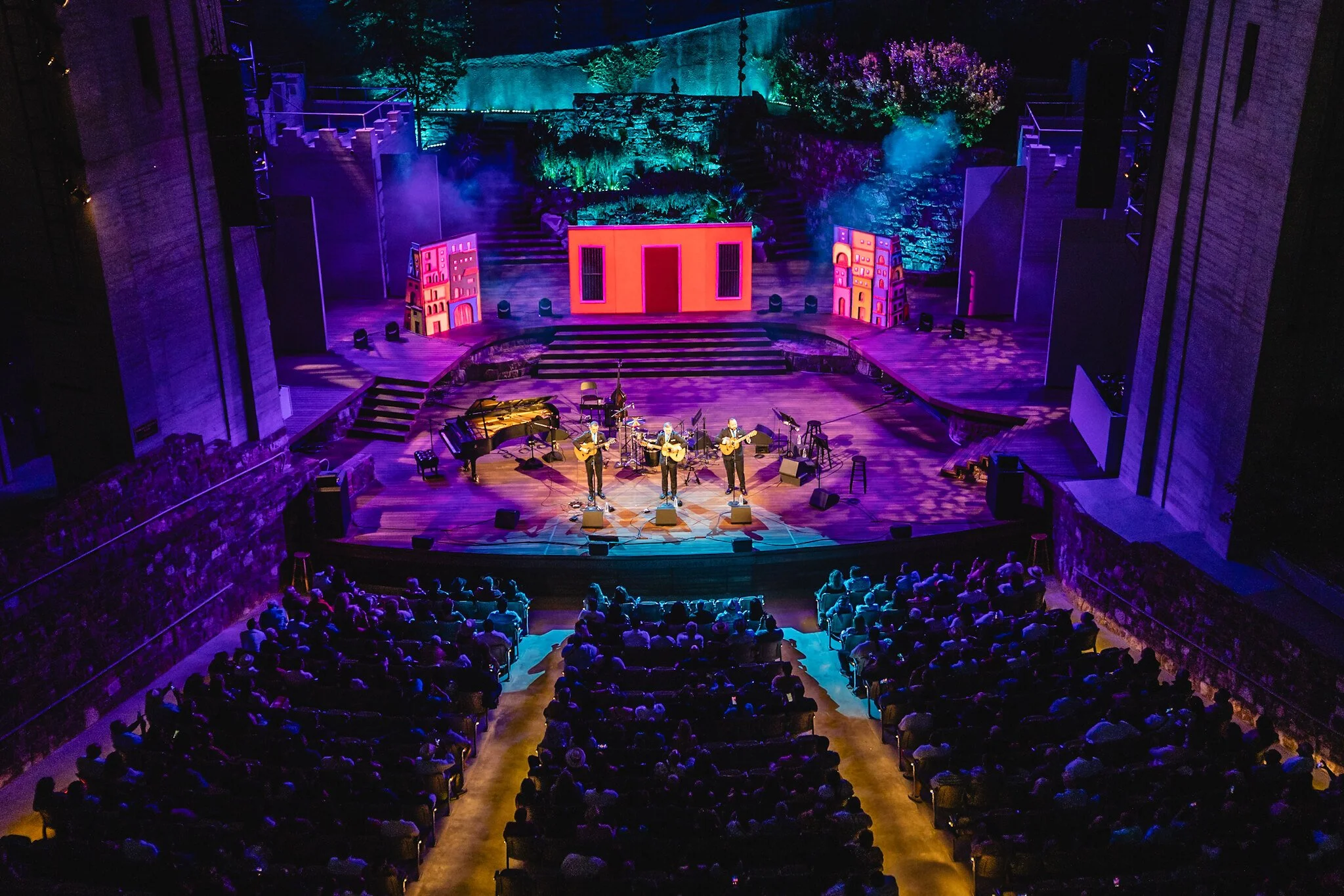 Live musical performance on a grand stage with three singers, a piano, and colorful lighting, attended by a large audience in an indoor concert hall.