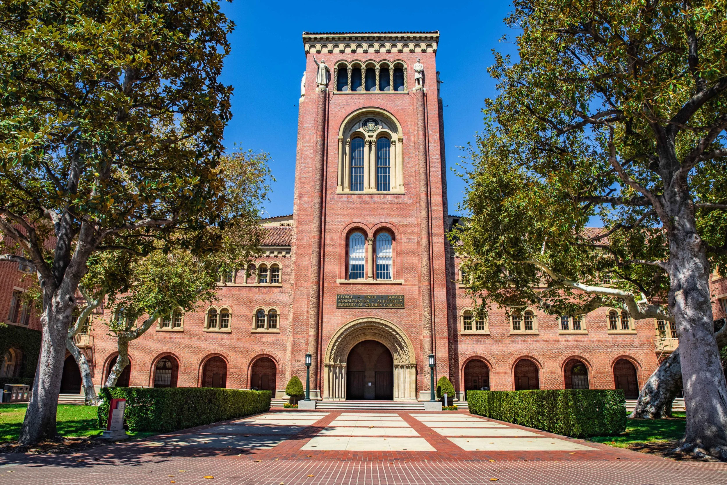 Red brick university building with arched entrance, tall tower, and Gothic-style windows, surrounded by trimmed bushes and trees under a blue sky.