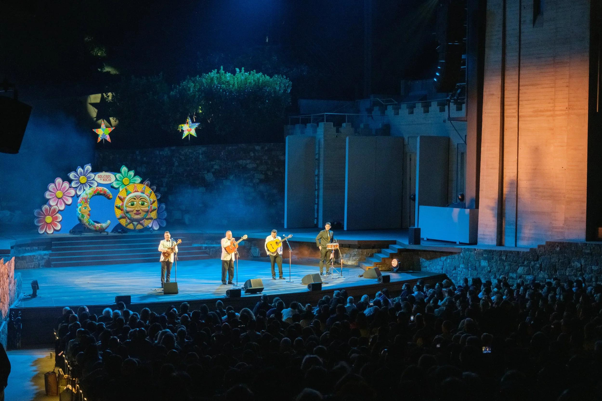 A live musical performance on a stage with four musicians playing guitars and singing, audience in foreground, colorful decorations including large flowers, a smiling sun, and crescent moon on the stage backdrop.