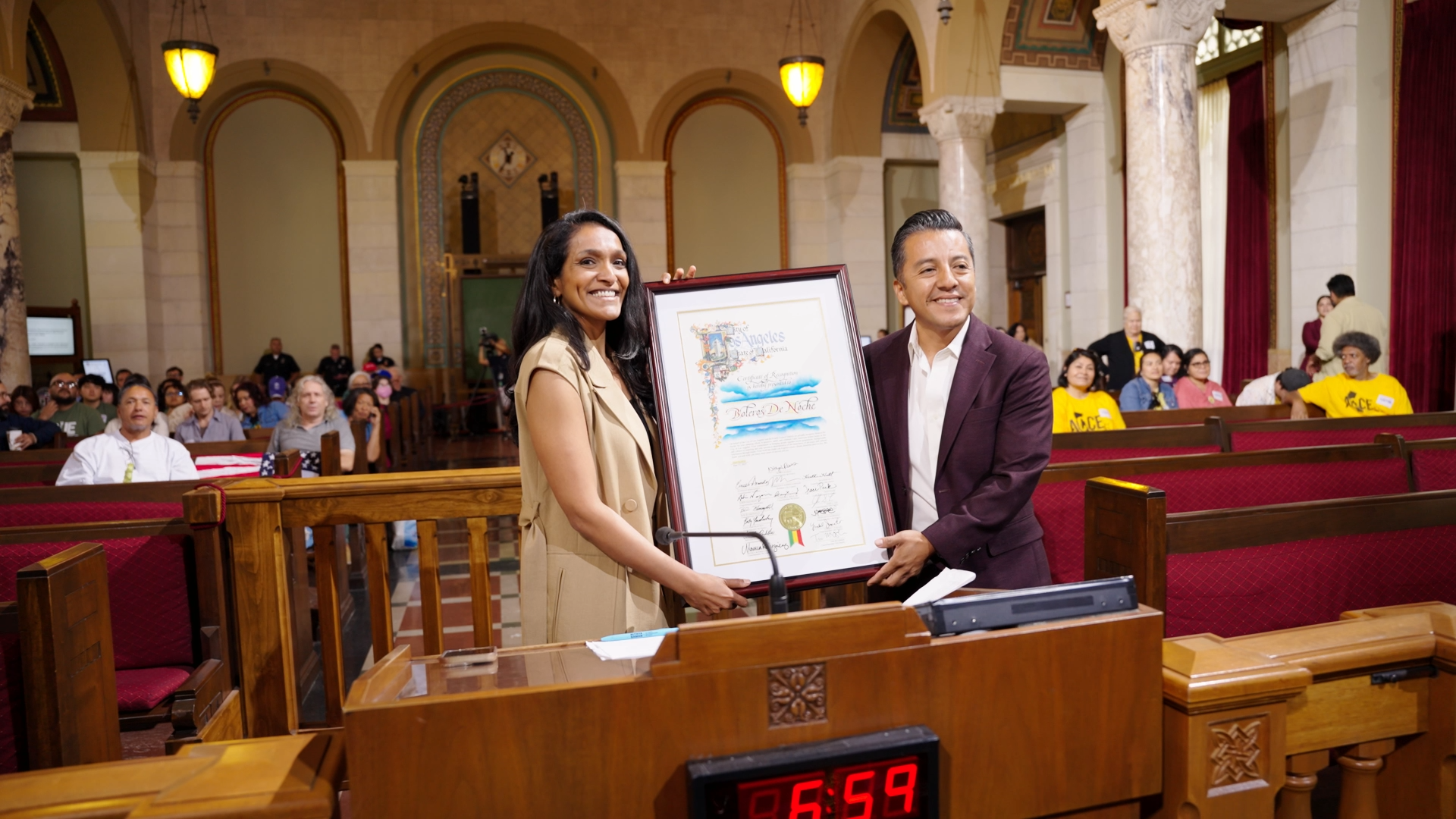A woman and a man smiling while holding a framed certificate or award in a large, ornate room with wooden furnishings and an audience watching.