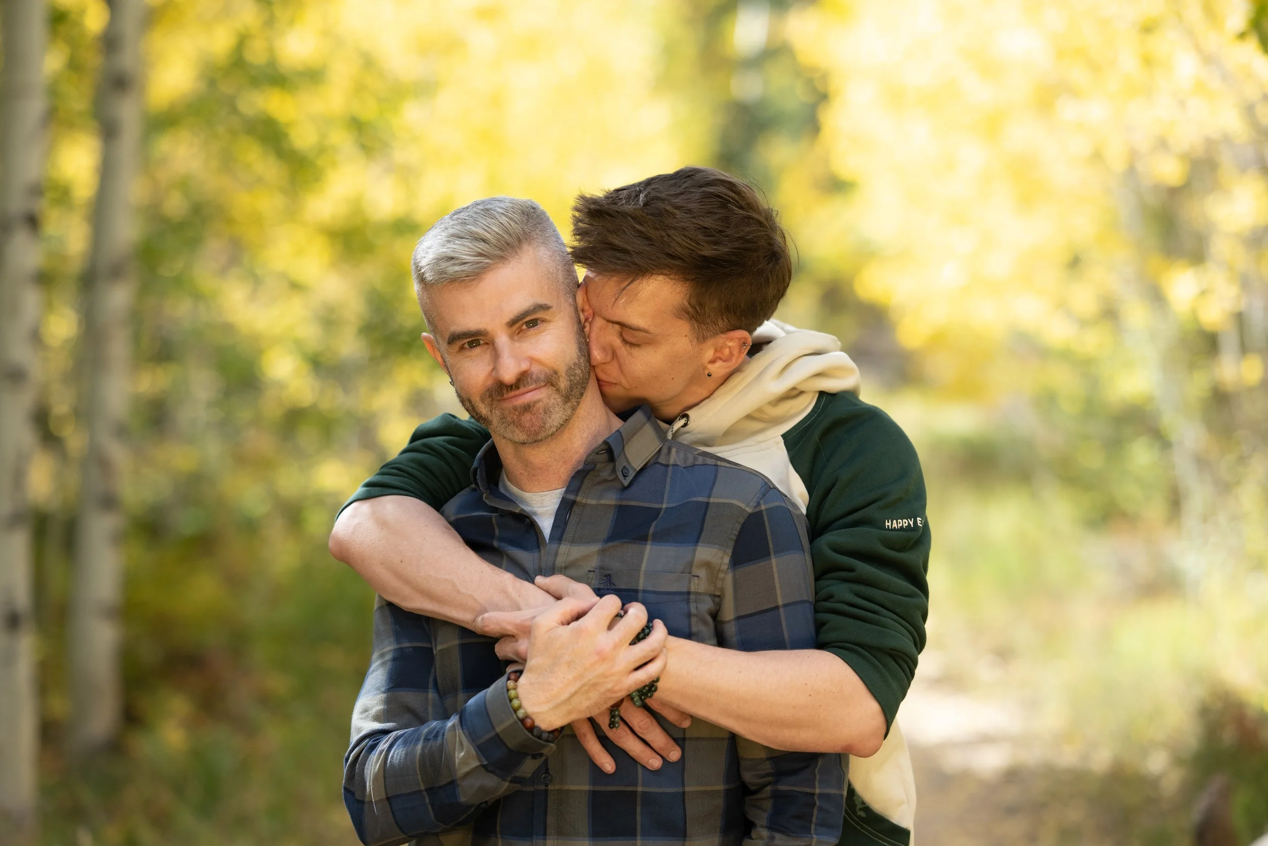 Two men embracing outdoors with autumn trees in the background, one kissing the other's cheek