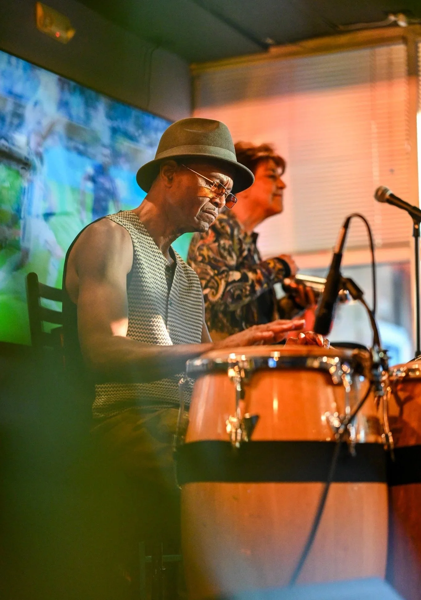 Two musicians performing indoors, one playing conga drums and the other playing a keyboard or similar instrument, with a colorful digital background and warm lighting.