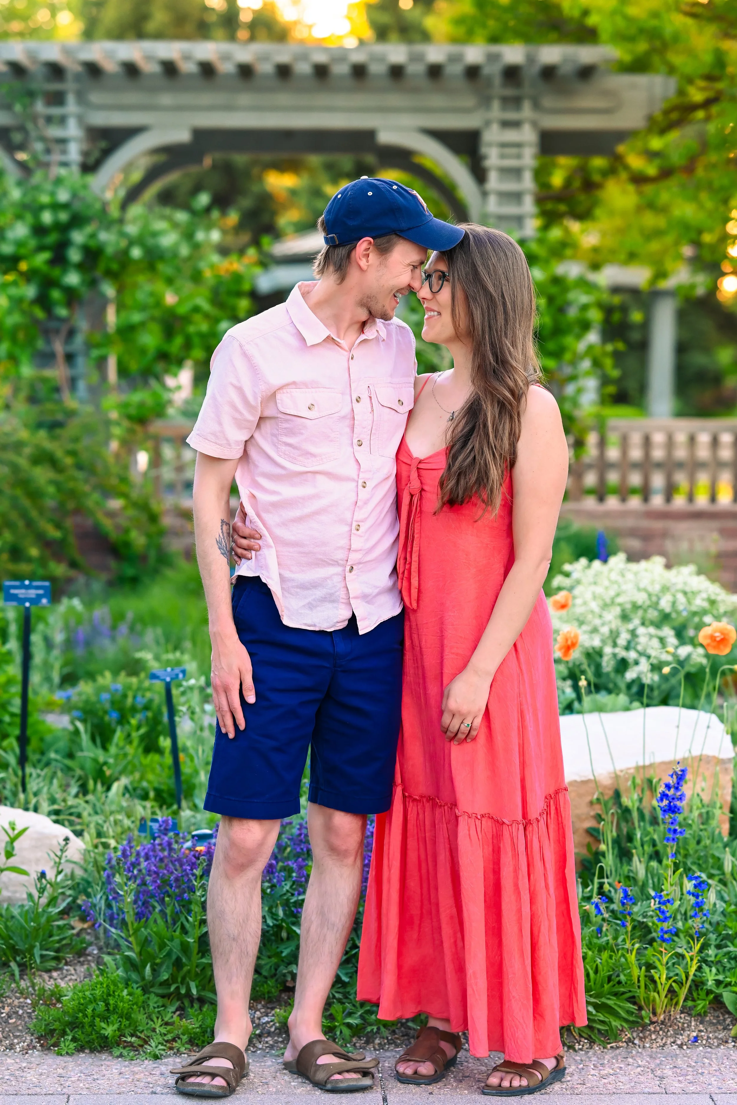 A young couple stands closely in a garden, smiling and about to kiss, with a wooden bridge and lush greenery in the background.