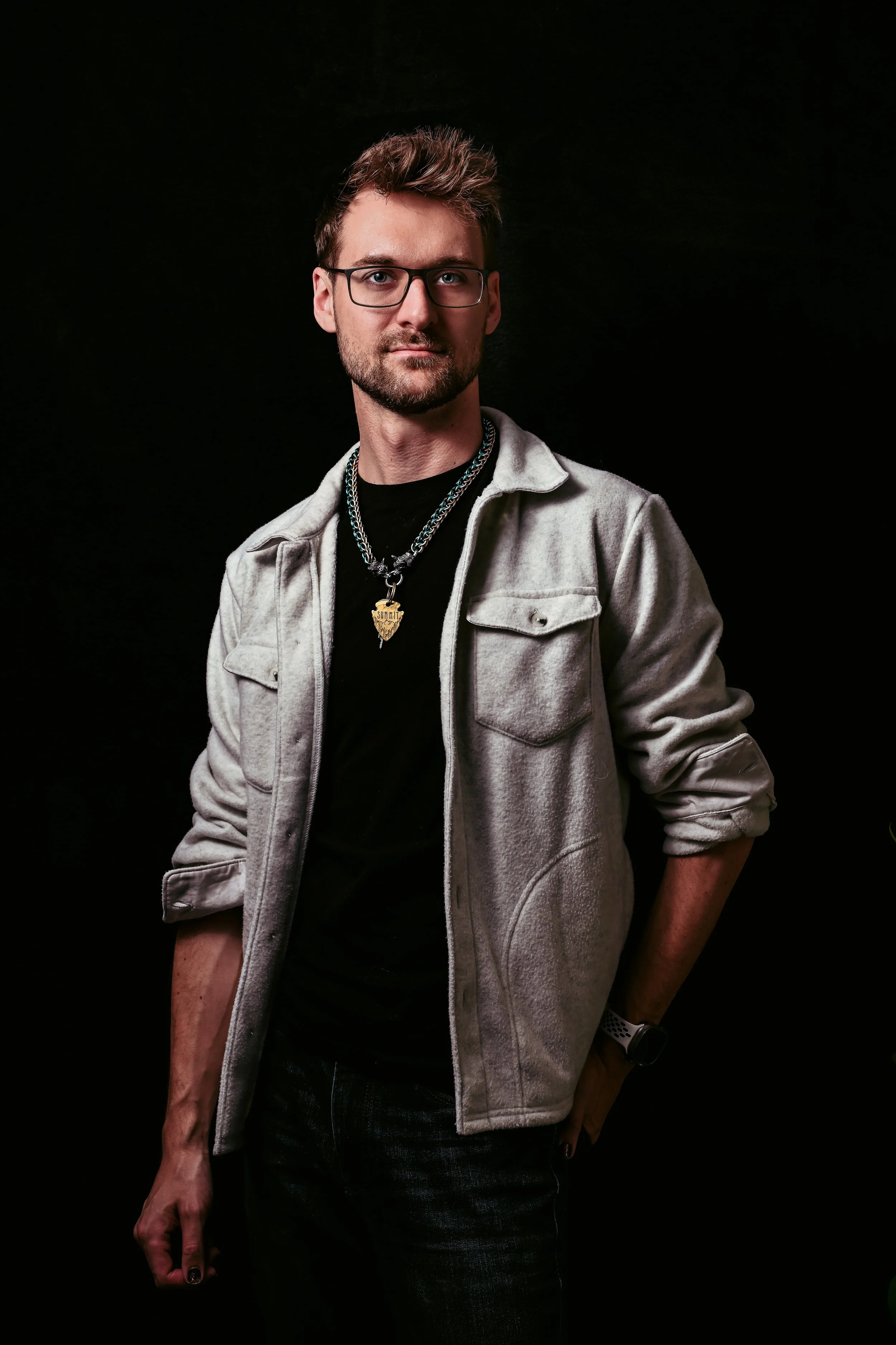 A young man with glasses and a beard, wearing a grey jacket, black shirt, layered necklaces, and a watch, standing against a dark background.