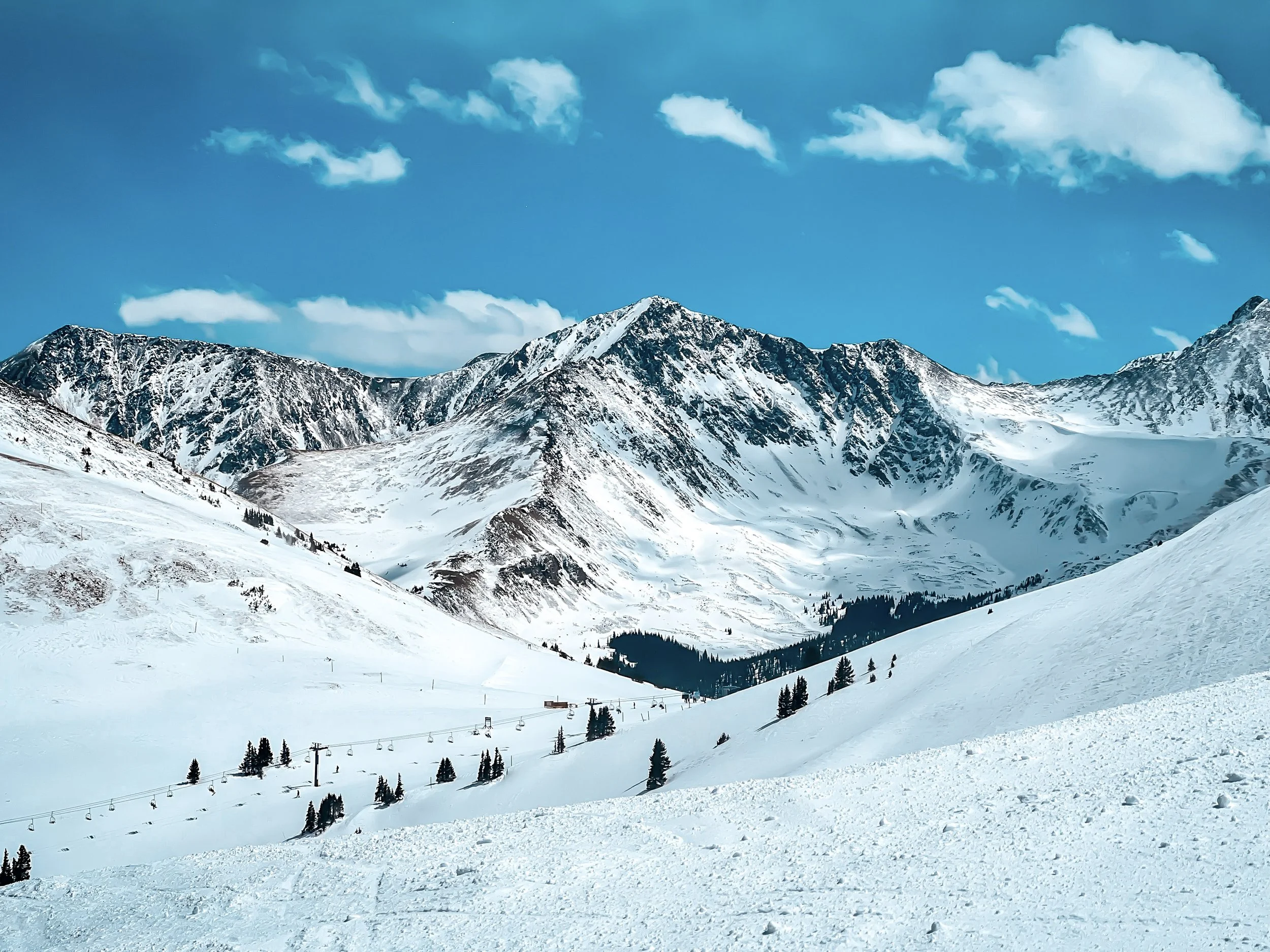 Snow-covered mountain range under a blue sky with scattered clouds, featuring ski lifts and pine trees in the foreground.