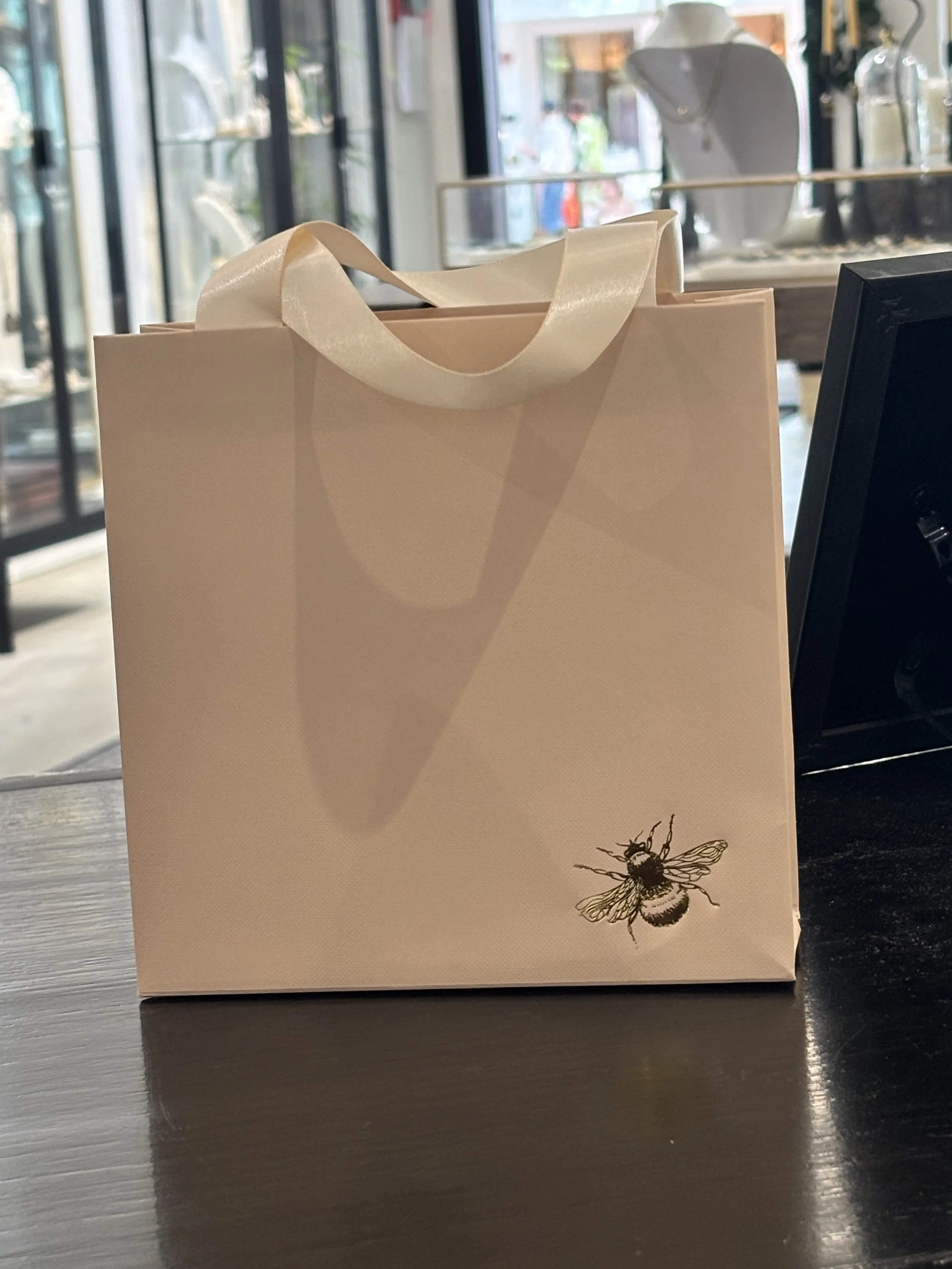 A beige shopping bag with cream-colored satin ribbon handles sitting on a dark wooden surface inside a shop or cafe. There is a detailed illustration of a bee in the bottom right corner of the bag. In the background, large windows let in natural light and show people walking outside.