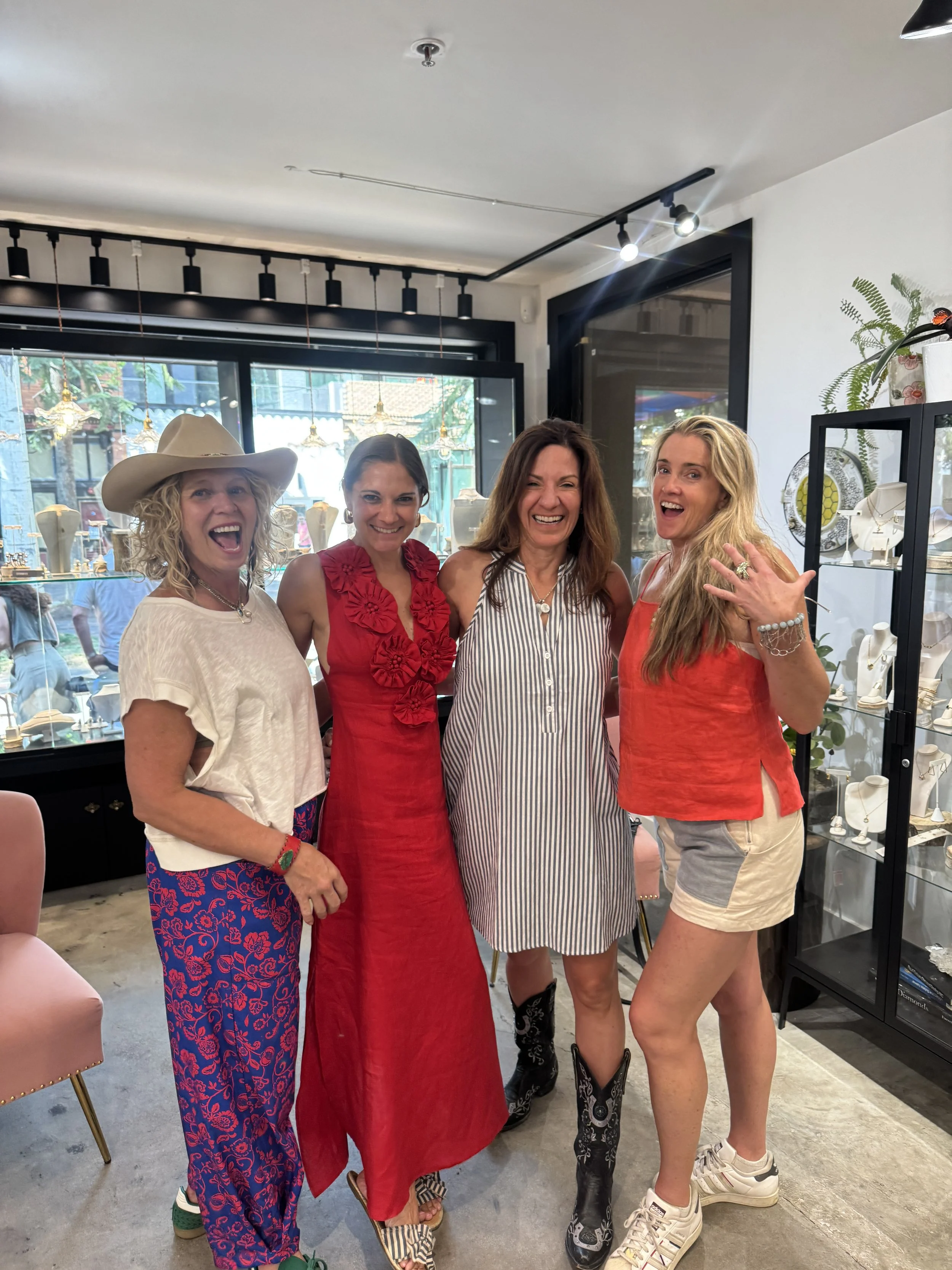 Four women standing together inside a jewelry store, smiling and posing for a photo. They are dressed in casual and colorful clothing, standing in front of display cases filled with jewelry.