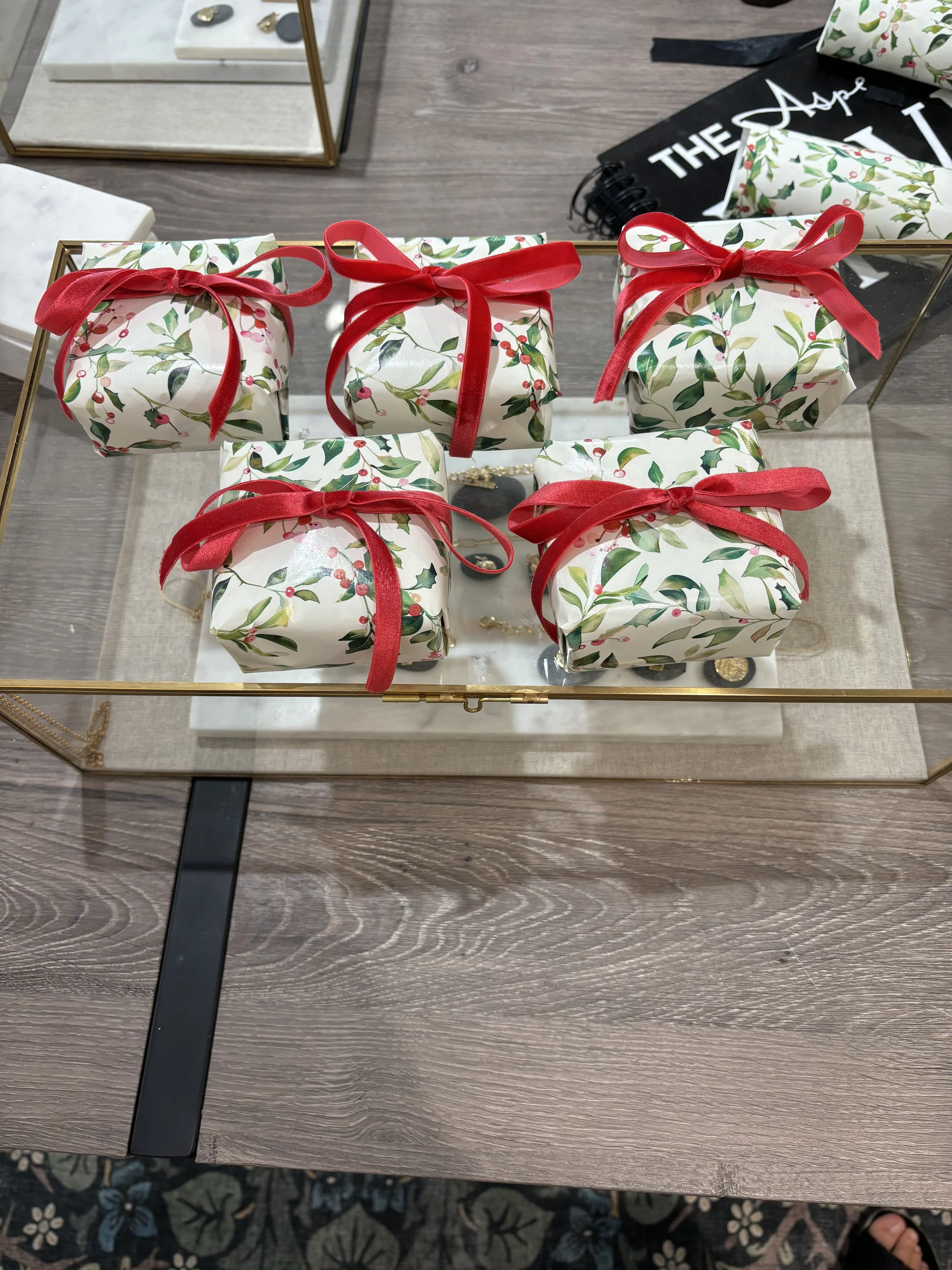 Six wrapped gifts with holiday-themed wrapping paper featuring holly and berries, tied with red ribbons, on a glass tabletop.