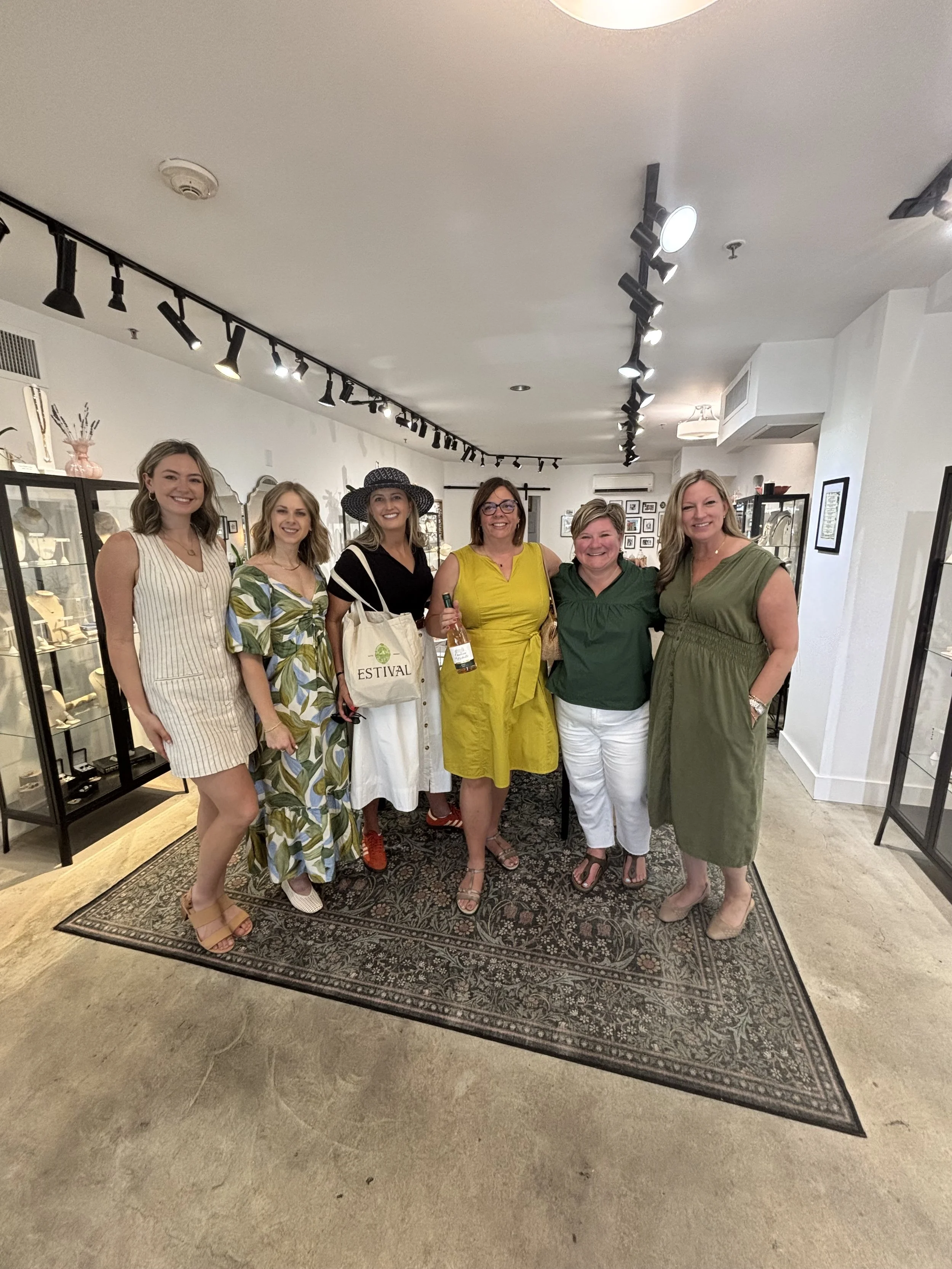 Six women standing indoors in a jewelry store, smiling at the camera. They are dressed in summer clothing, with one woman holding a bottle of wine and a paper bag with the word "ESTIVAL". The store has display cabinets with jewelry and artwork on the walls, under black track lighting.