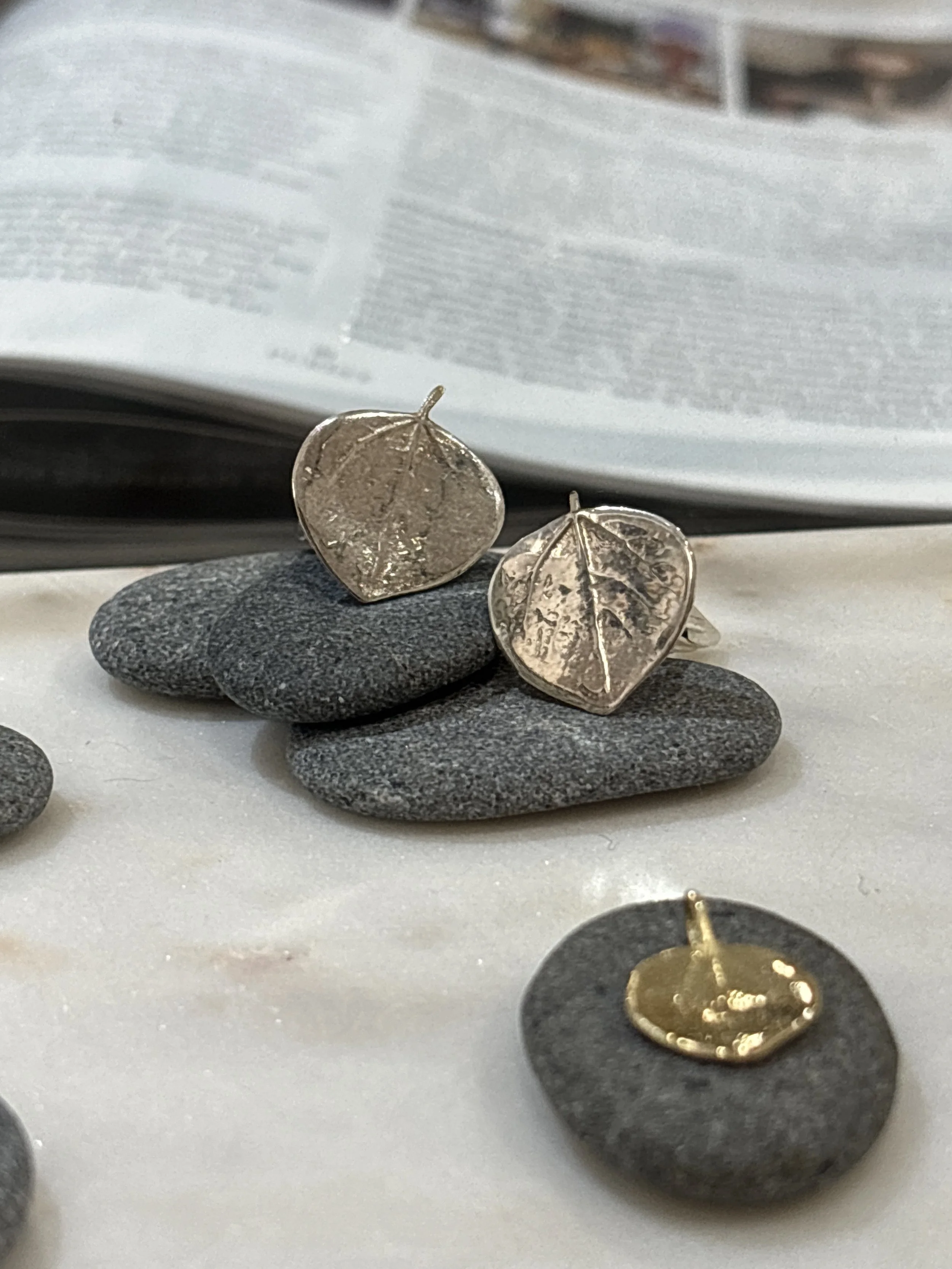 Close-up of a pair of silver leaf-shaped earrings resting on black stones, with jewelry display in the background.