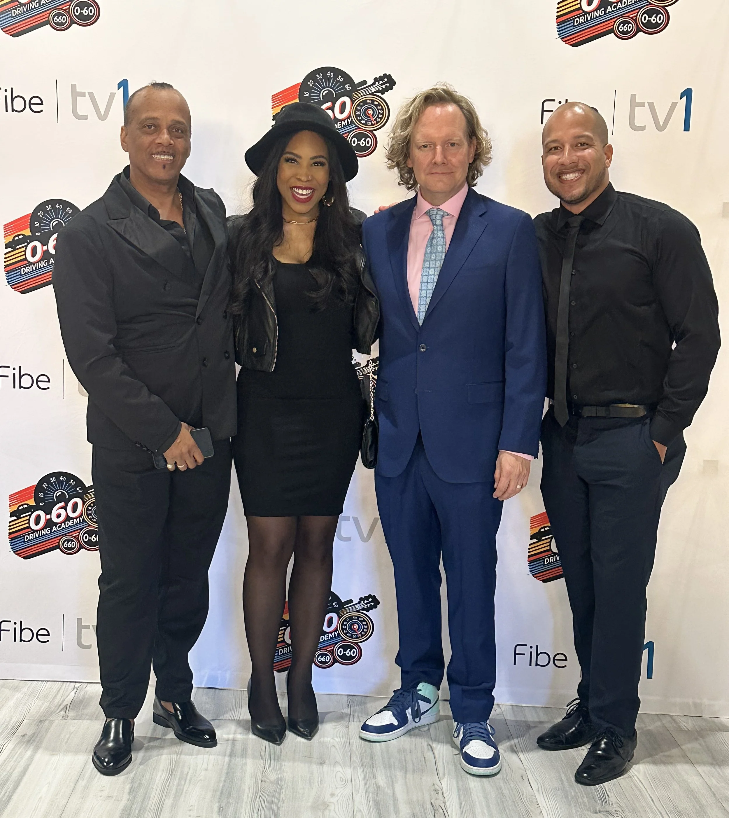 Four people standing in front of a white backdrop with logos. The group includes two men and one woman. The woman is wearing a black hat, black dress, and leather jacket. The man next to her is wearing a blue suit, pink shirt, and patterned tie. The 