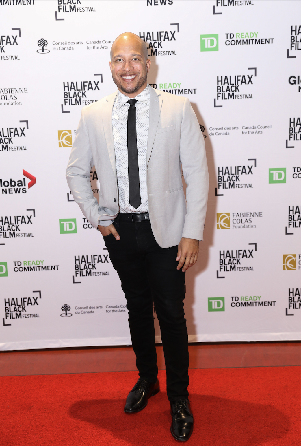 A man with a shaved head and a beard, wearing a light-colored blazer, white shirt with a subtle pattern, black tie, black pants, and black shoes, standing on a red carpet at the Halifax Black Film Festival. The background features a white promotional
