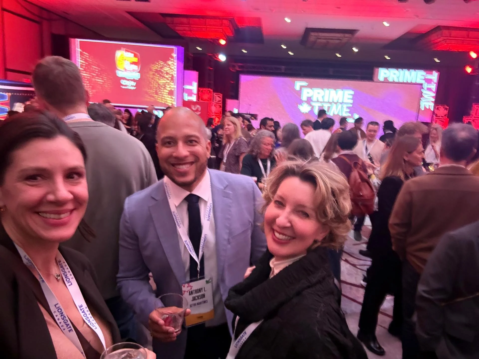 A group of people at a conference or event, smiling and posing for a photo. The background shows a large screen with the words 'PRIME TIME' and a crowd of attendees.