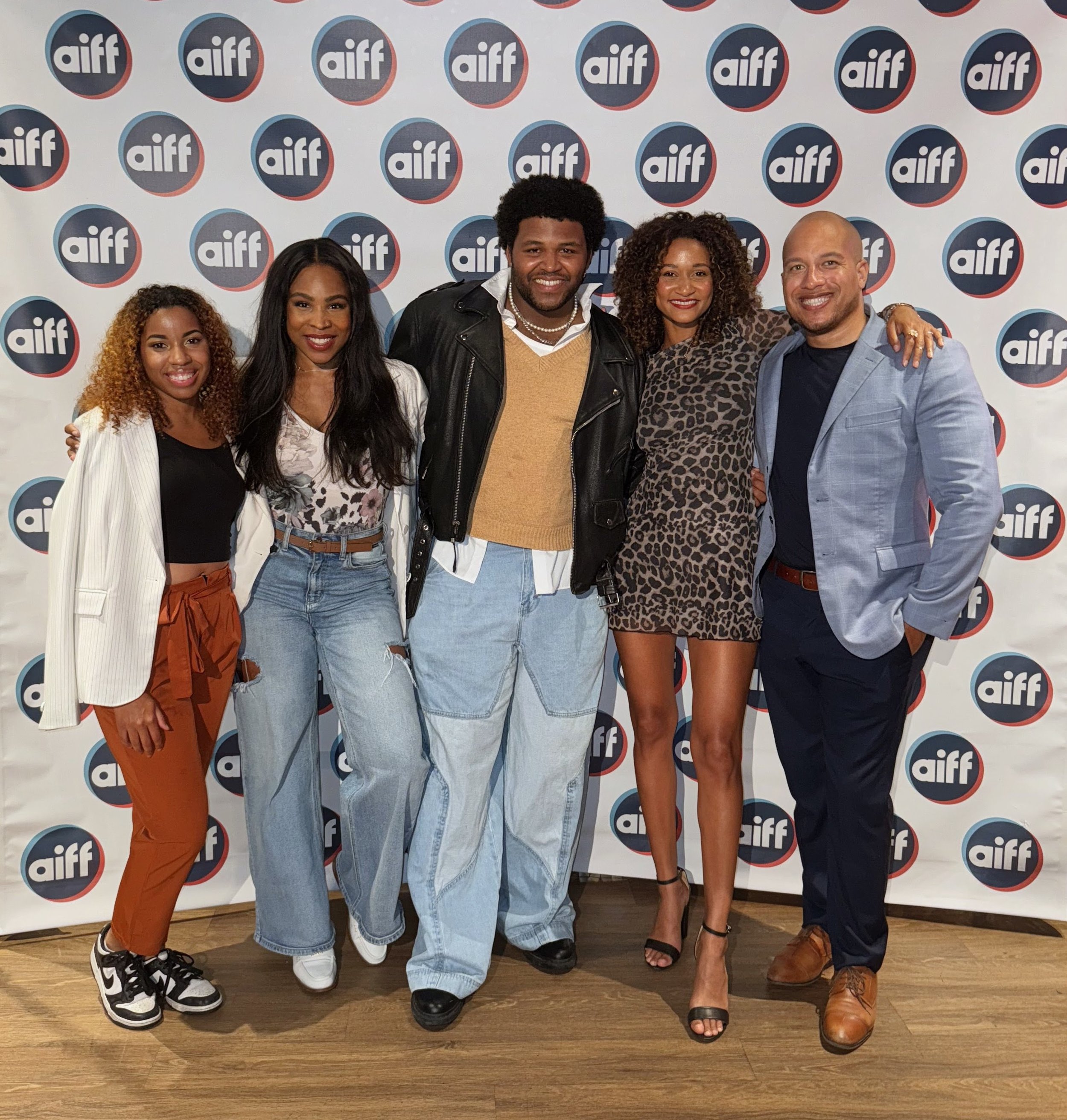Six diverse individuals posing in front of a step and repeat backdrop with the 'aiff' logo at the Austin International Film Festival. The group includes four women and two men, dressed in stylish, casual to semi-formal clothing, smiling and standing 