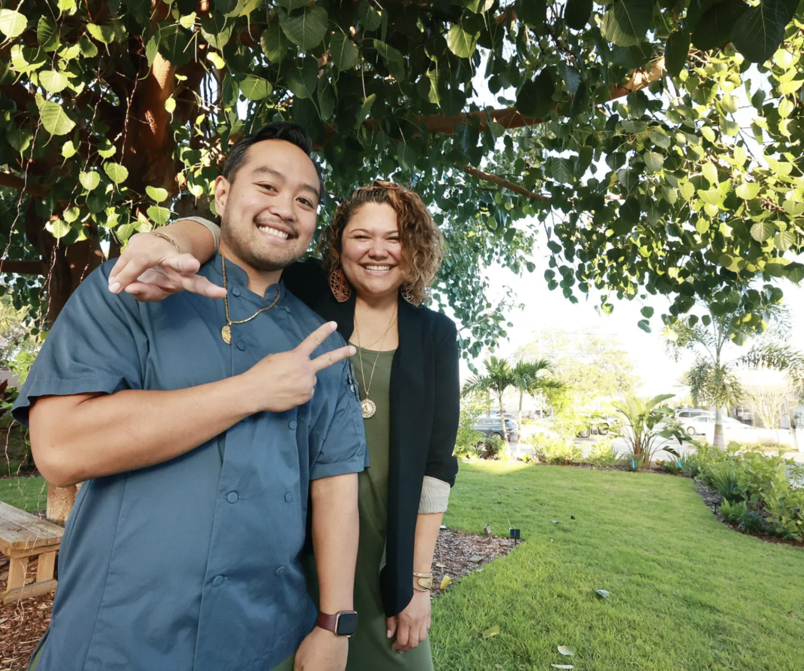 Two people smiling under a tree, with one person making a peace sign, in a garden setting.