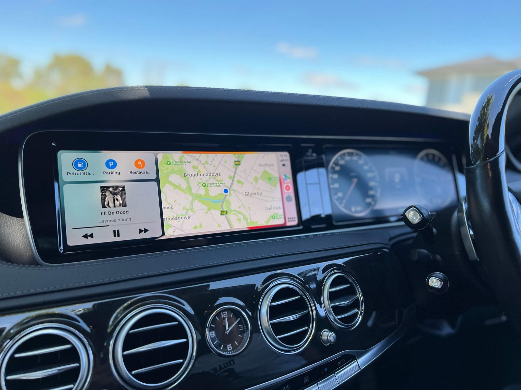 Car interior dashboard showing a digital display with navigation map, music player, and various controls, with a view of blue sky outside.