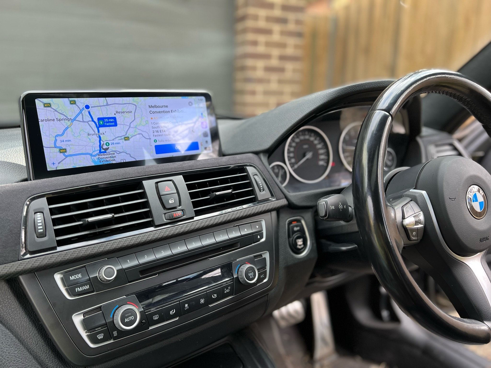 Inside a BMW vehicle, the dashboard shows navigation on a screen indicating travel routes to the Melbourne Convention Exhibition Center, with controls for heating and audio system below.