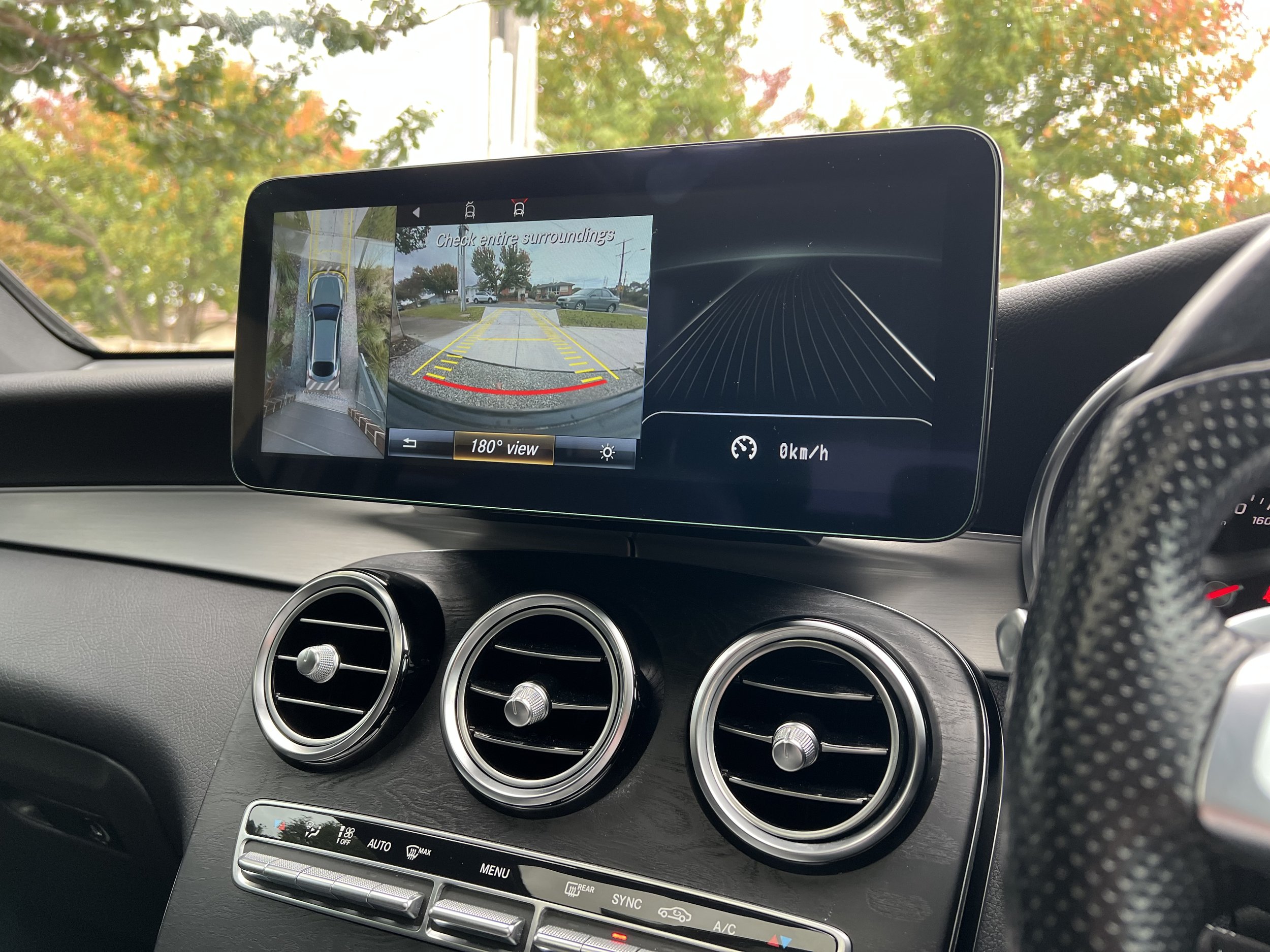 Car's dashboard showing a 360-degree camera display with rear view and a black navigation screen, surrounded by black air vents and climate control buttons, outside view of trees with fall foliage.