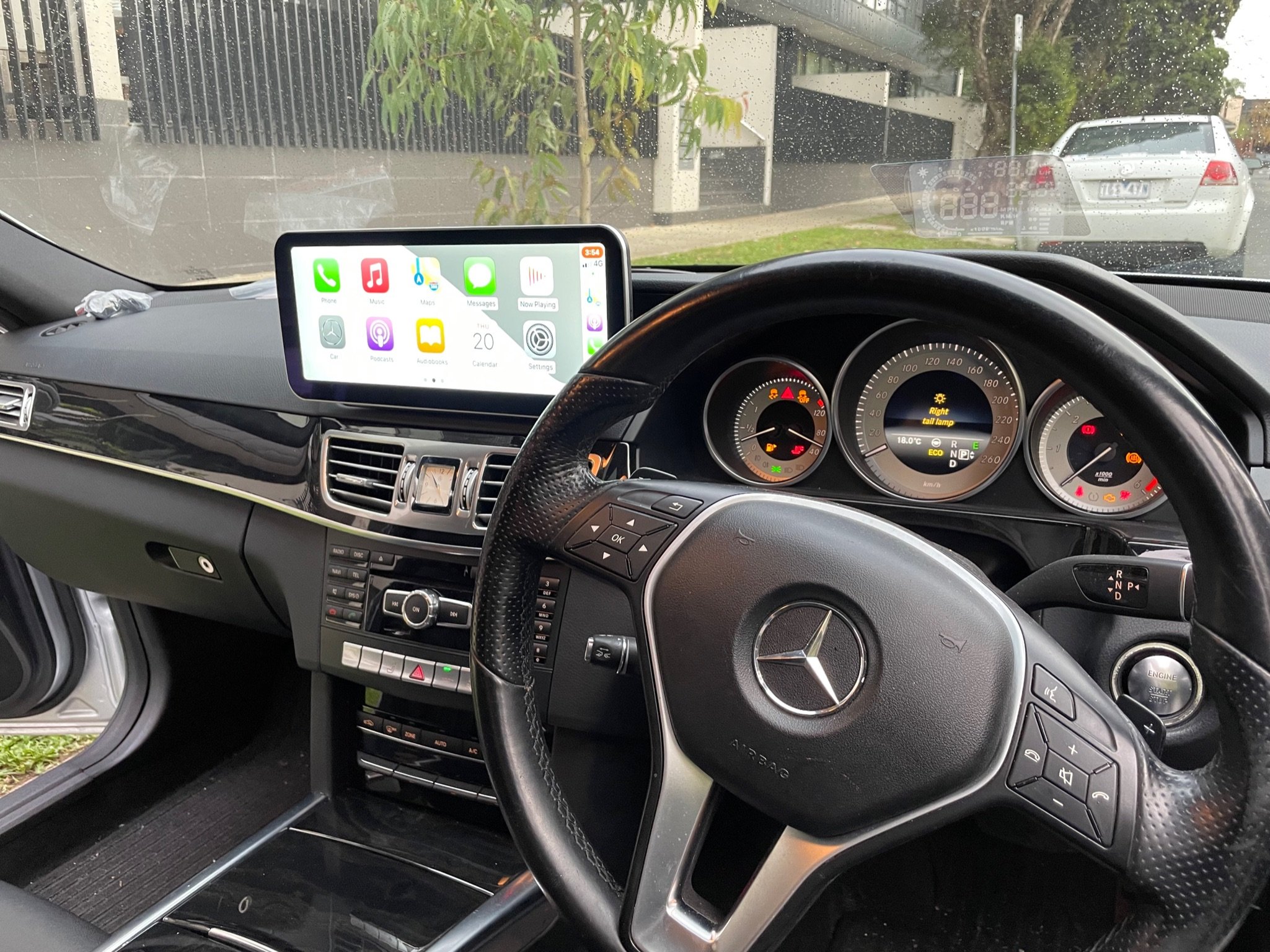 Inside view of a Mercedes-Benz car dashboard with a digital display, GPS screen, and steering wheel in focus, parked on a street with other vehicles visible outside.