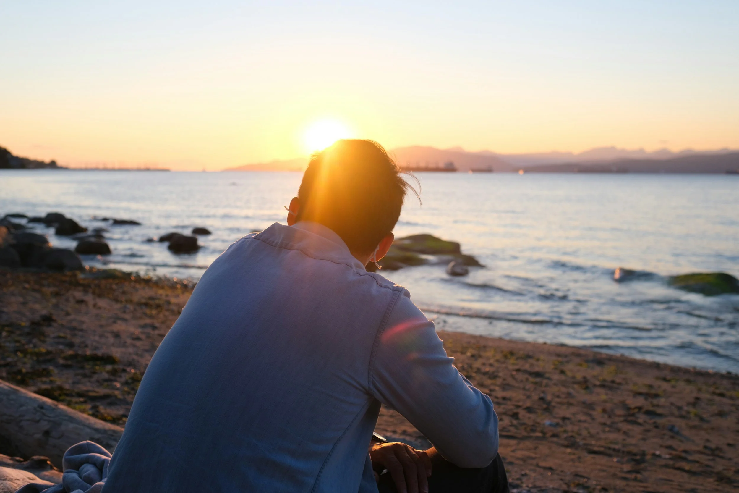 Person sitting on a rocky beach facing the water and a sunset, with the sun near the horizon and mountains in the distance.