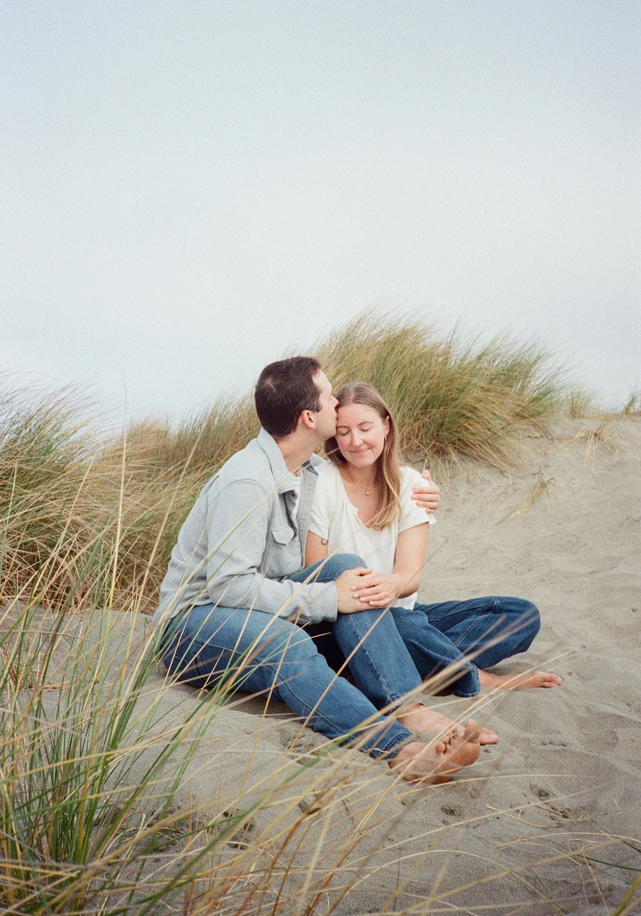 A young couple sitting on a sandy beach with tall grass, the man giving the woman a kiss on her forehead, both with their eyes closed and smiling, wearing casual clothes.