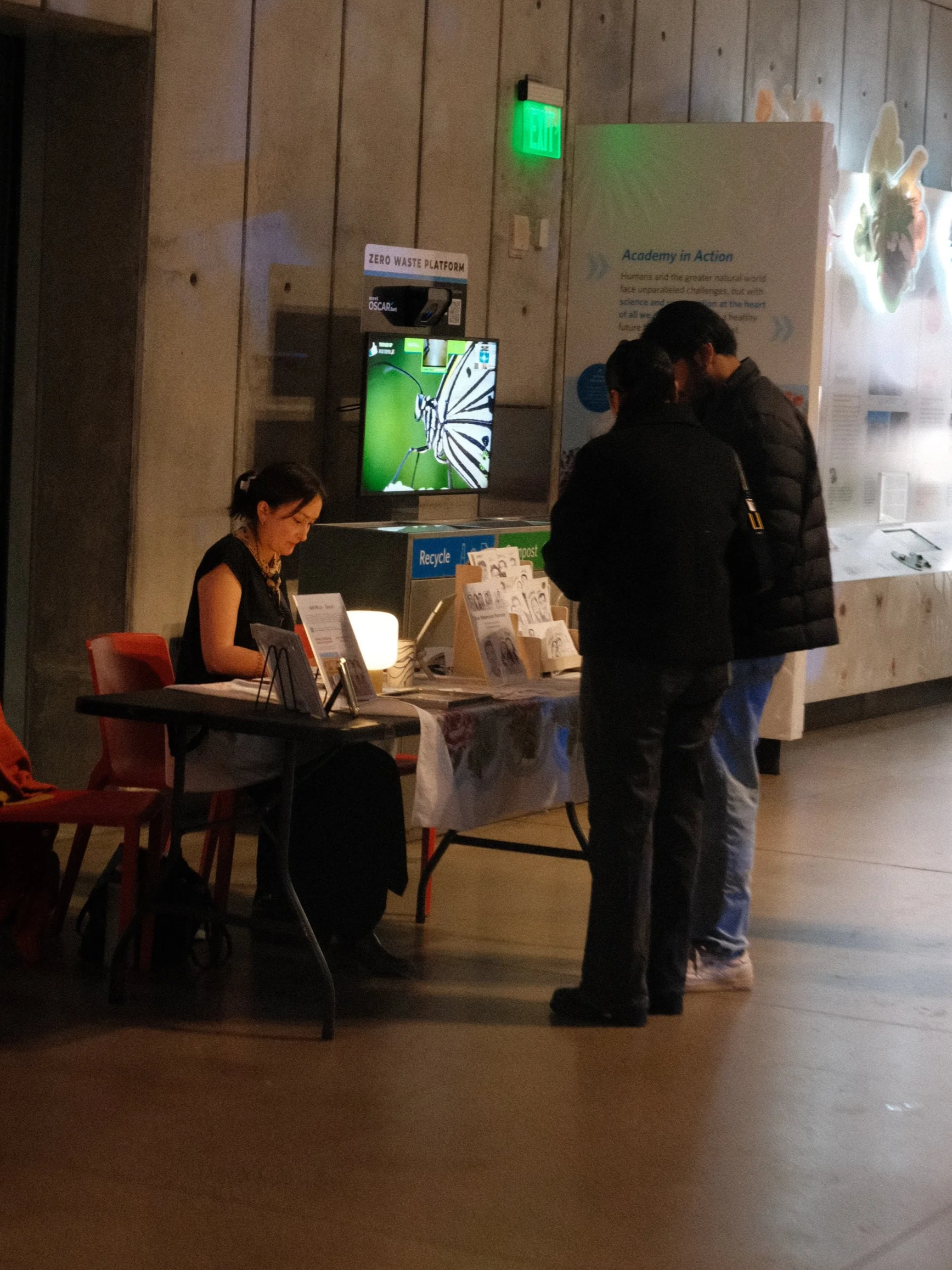 People standing at a table with informational materials at an indoor event, with a monitor displaying a butterfly, and a sign that reads 'ZERO WASTE PLATFORM' in the background.
