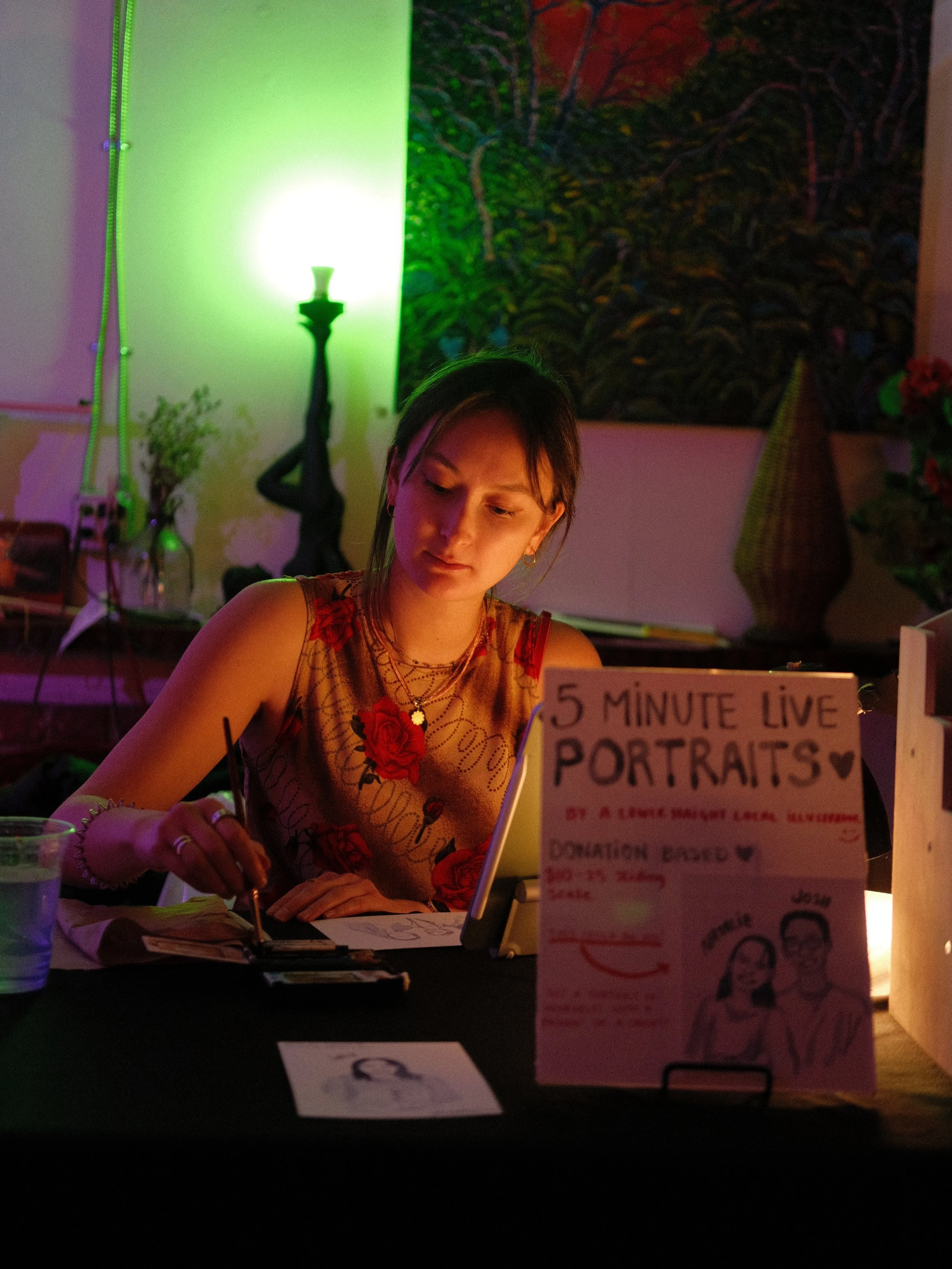 A young woman sitting at a table during a live portrait sketch event, with a sign advertising '5 Minute Live Portraits'. The setting has colorful lighting, a painting on the wall, and various items on the table.