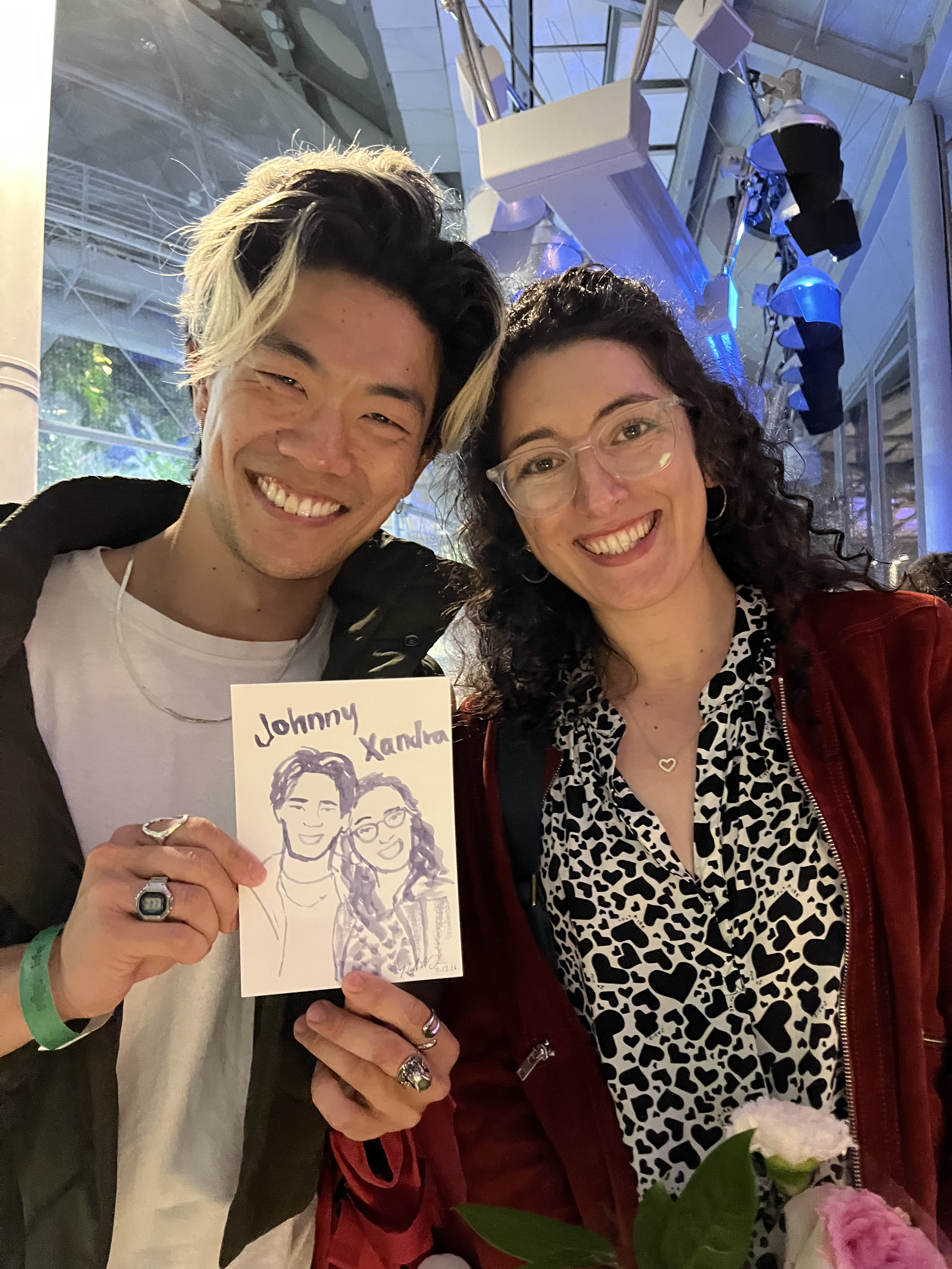 A man and woman smiling for a photo indoors, with the man holding a drawing of both of them labeled 'Johnny' and 'Xandra'. They are wearing casual clothing, and there are flowers in the foreground.