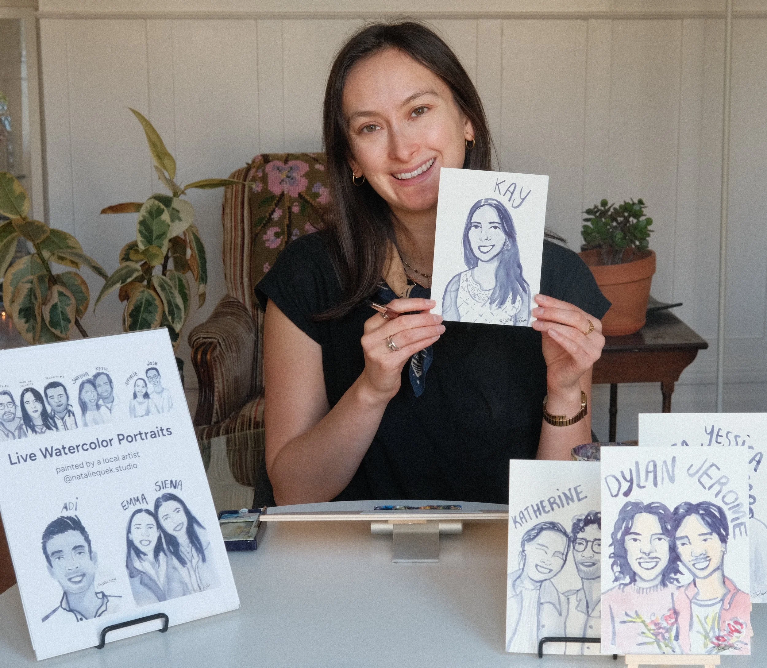 Woman smiling at a watercolor portrait she created, holding the drawing of herself labeled 'Kay', with various other watercolor portraits of people displayed on the table.