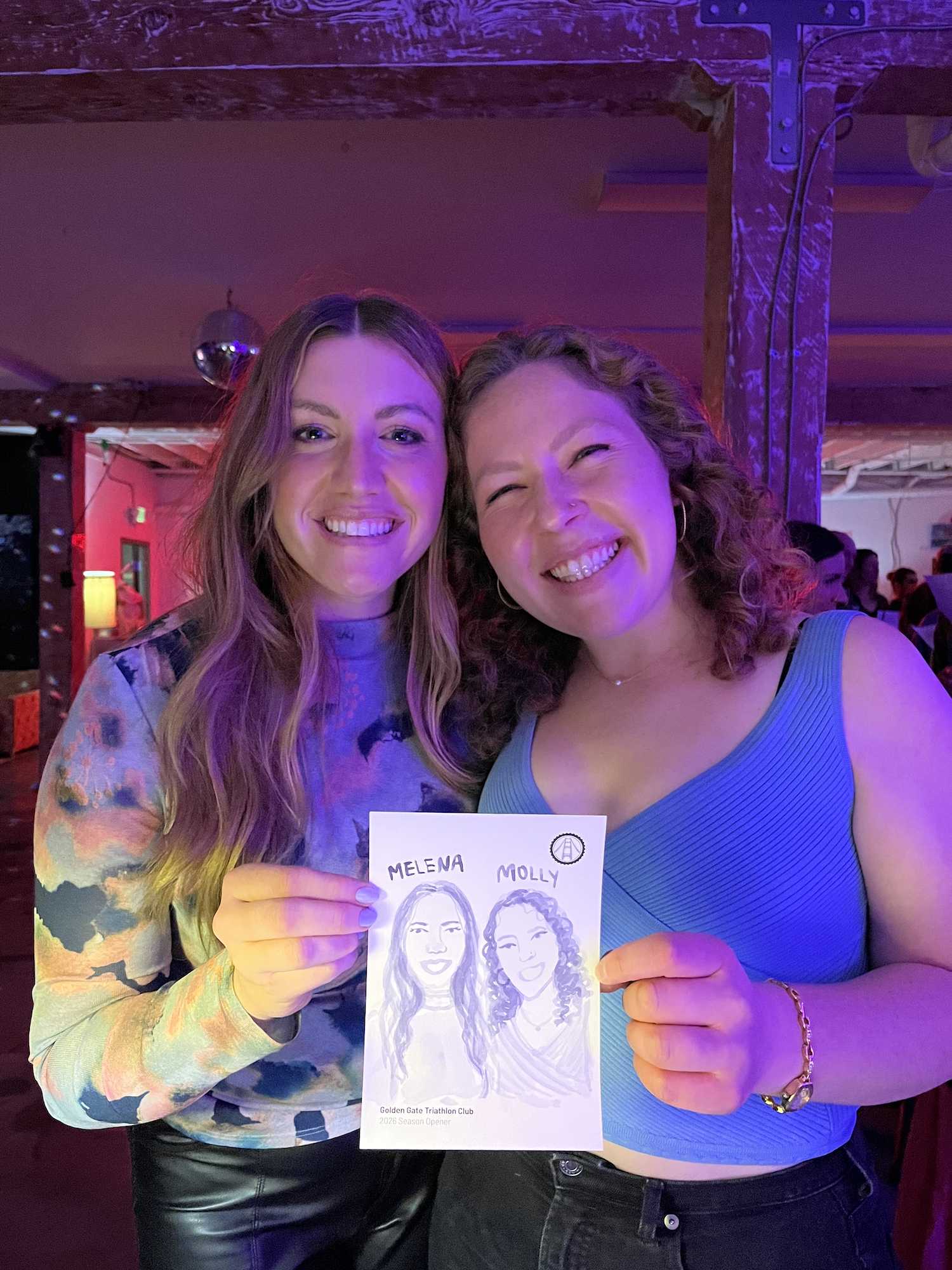 Two women smiling and holding a drawing of themselves with their names, Melena and Molly, on a piece of paper at a social event with purple lighting.
