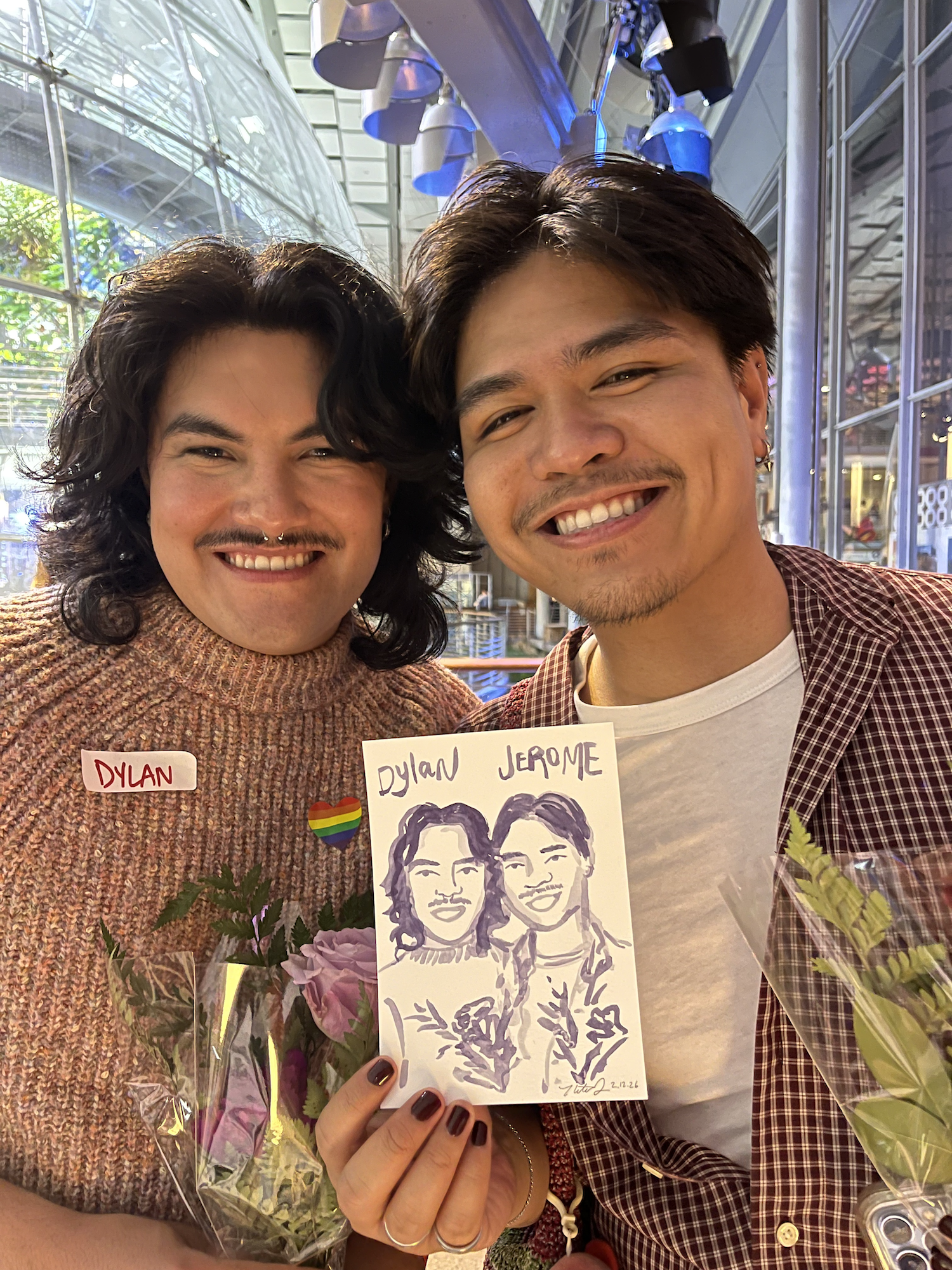 Two smiling individuals hold a sketch of themselves, in an indoor setting with glass walls and hanging blue lights, one wearing a rainbow heart pin and a salmon sweater, the other in a checkered shirt and earrings.