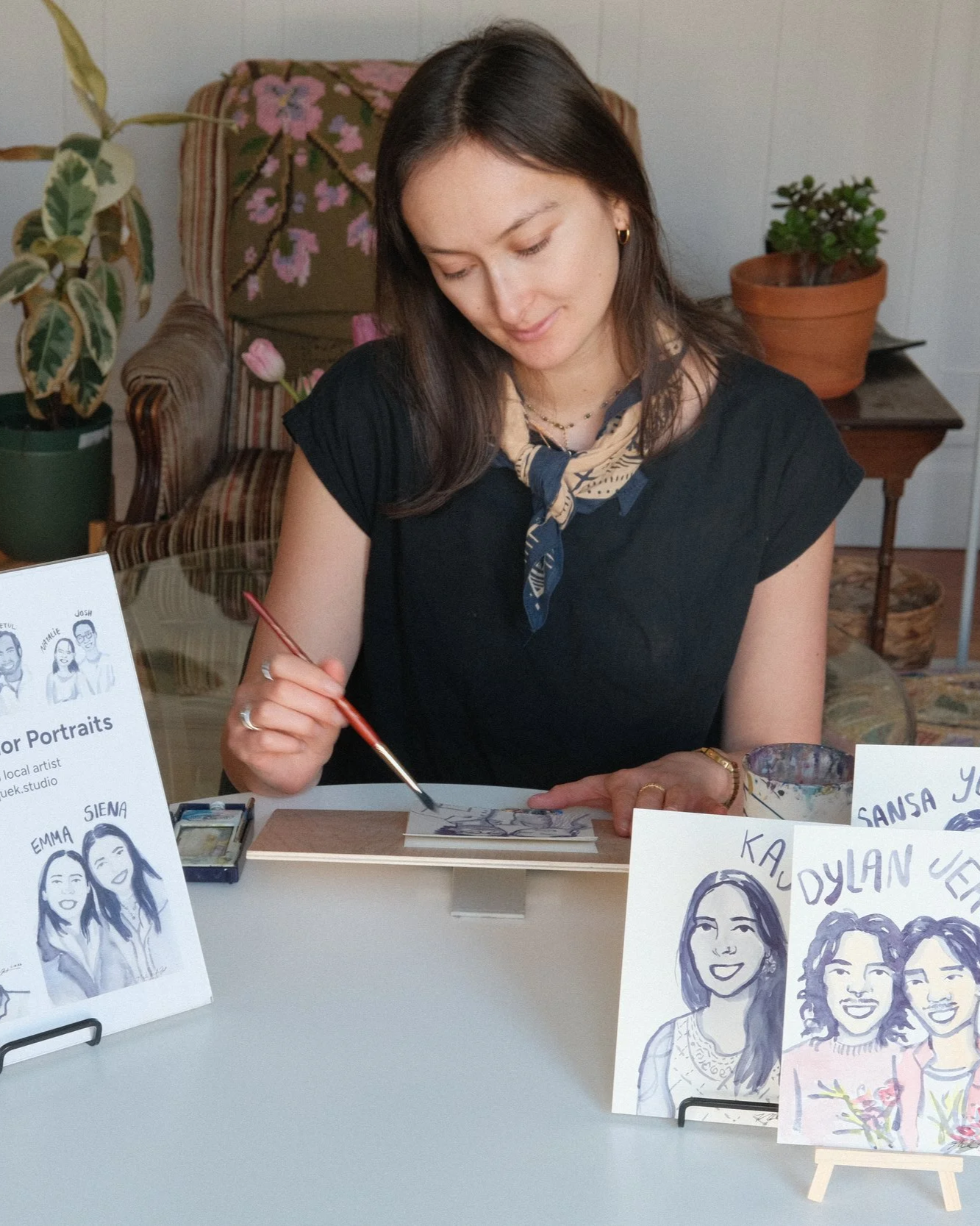 Woman painting watercolor portraits at a table with sample portraits displayed on stands.