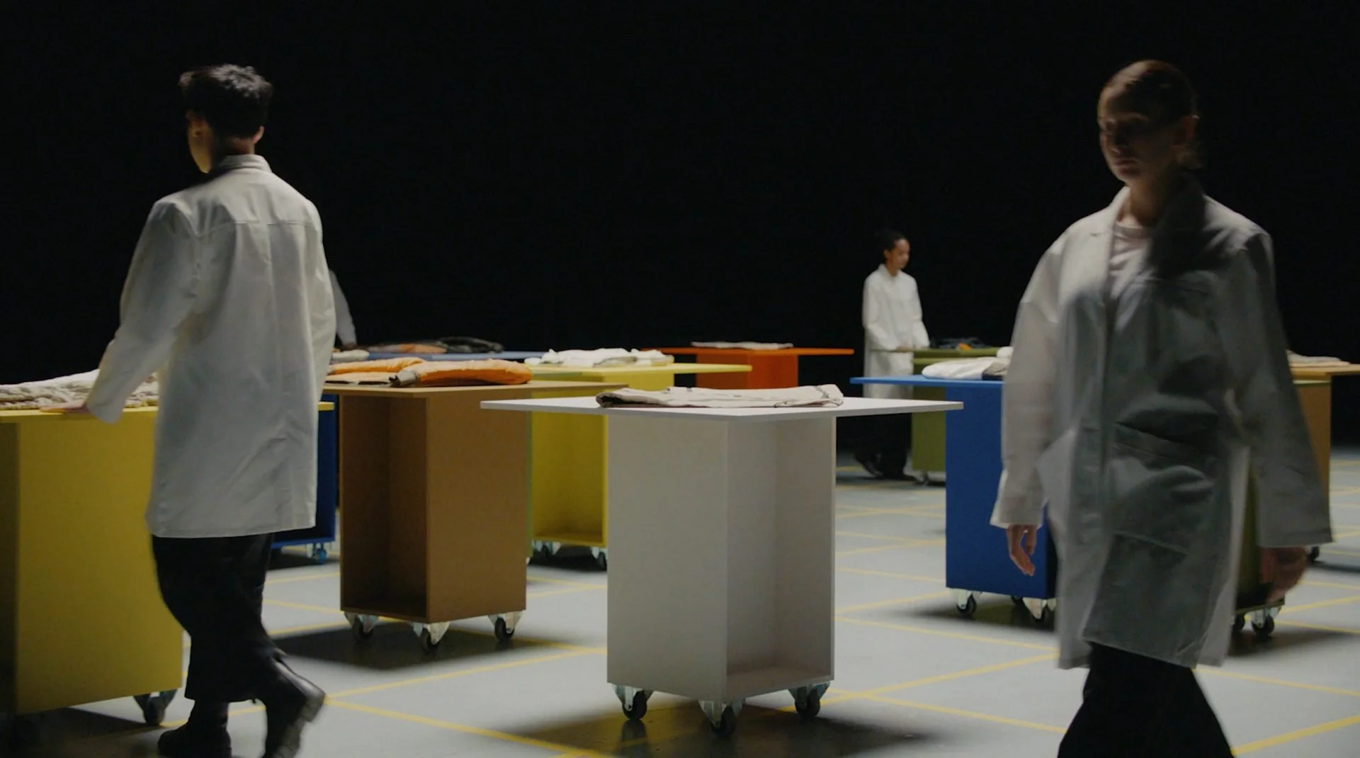 Marfa Stance film still: performers in white coats moving through colour-blocked plinth grid
with garments, DOP James Dougan