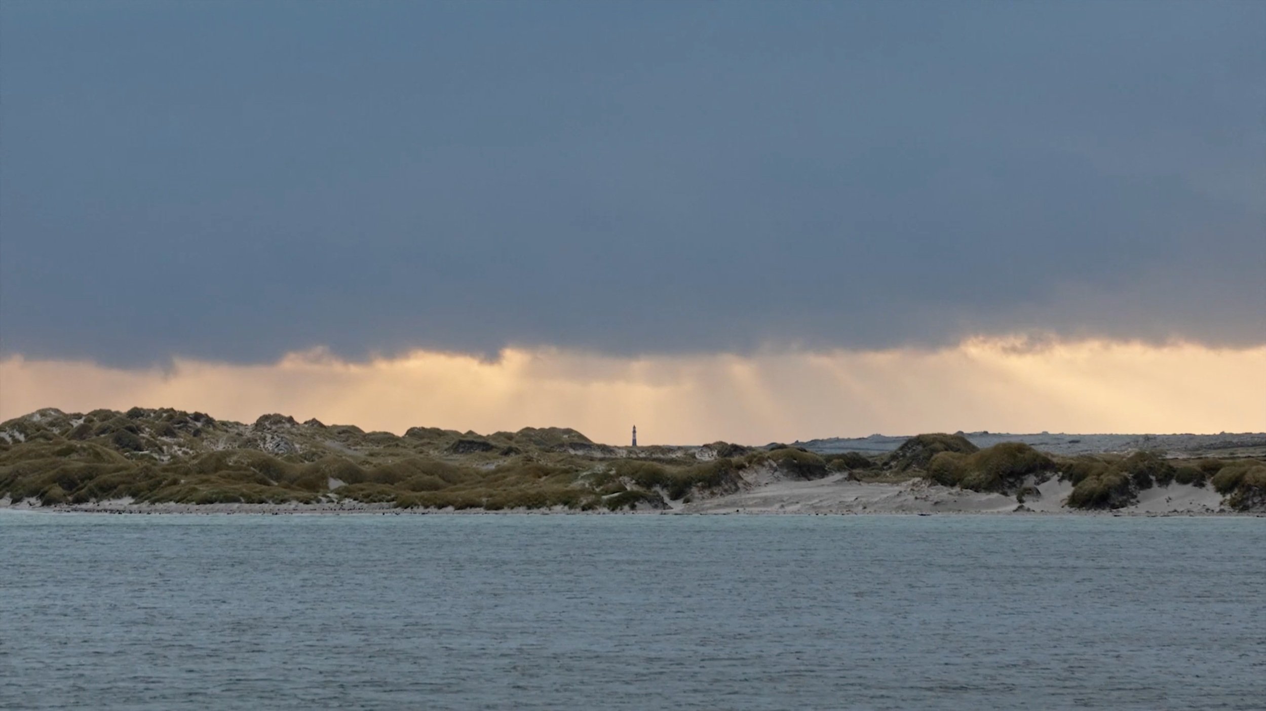Falkland Islands coastline under a stormy South Atlantic sky, looking across sheltered water toward distant shores — filmed for Connecting The Falklands, Square and Mastercard